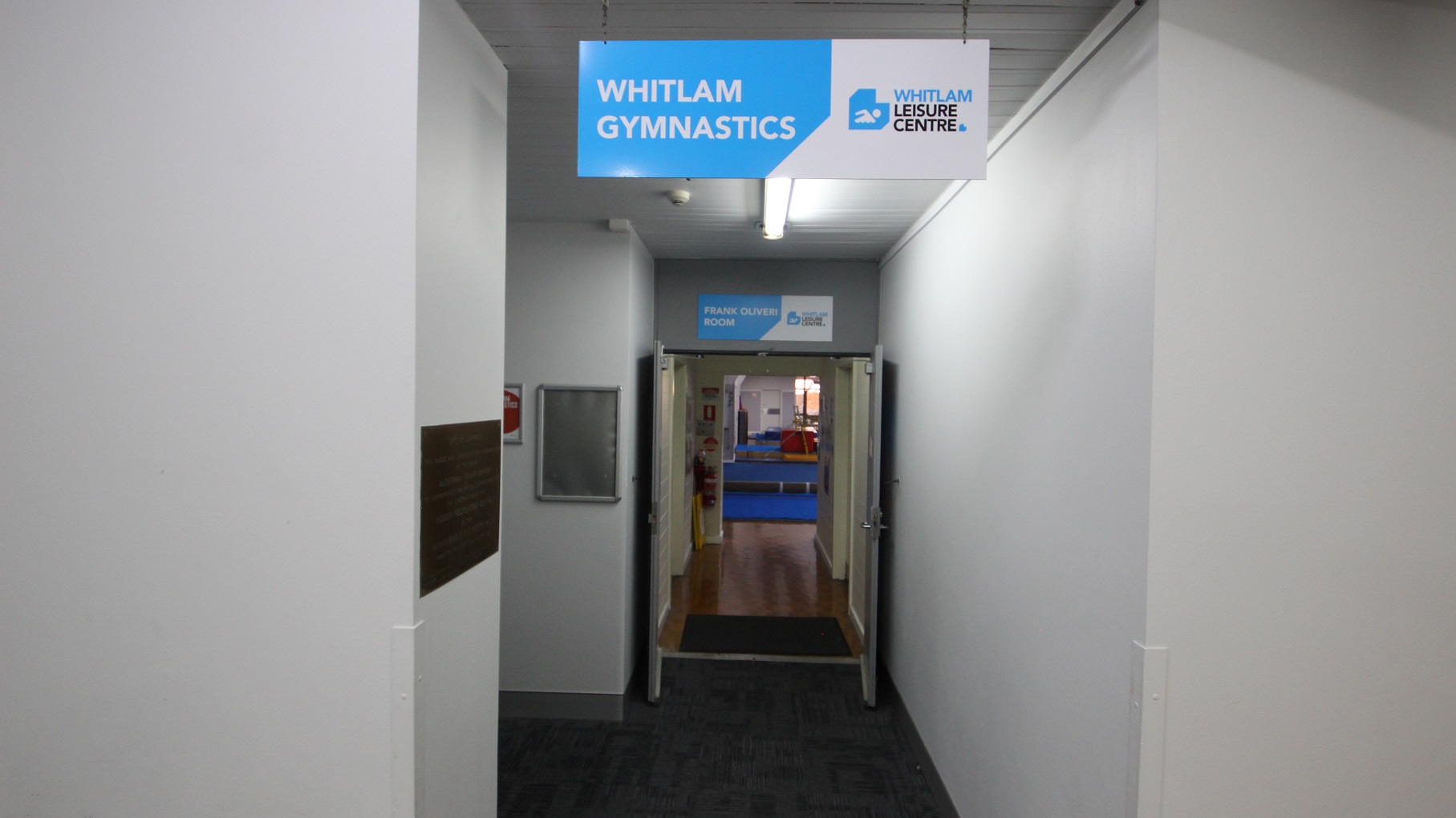 Corridor leading towards Whitlam Gymnastics. The walls are clean and are painted by white. Above the first open door is the signboard containing Frank Oliveri Room, and on the next door is a fire distinguisher mounted on the lower part of the wall. There are large blue mats inside the Gymnasium. Corridor leading towards Whitlam Gymnastics. The walls are clean and are painted by white. Above the first open door is the signboard containing Frank Oliveri Room, and on the next door is a fire distinguisher mounted on the lower part of the wall. There are large blue mats inside the Gymnasium.