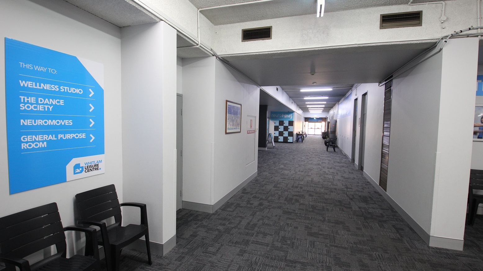 Corridor leading to the Wellness studio. Similar to the other floors of the different kinds of amenities inside the Whitlam Leisure Centre, this also has a grey tiled floor with zigzag lines as a design. A number of black chairs and locker cabinets are placed on the sides of this corridor. Corridor leading to the Wellness studio. Similar to the other floors of the different kinds of amenities inside the Whitlam Leisure Centre, this also has a grey tiled floor with zigzag lines as a design. A number of black chairs and locker cabinets are placed on the sides of this corridor.