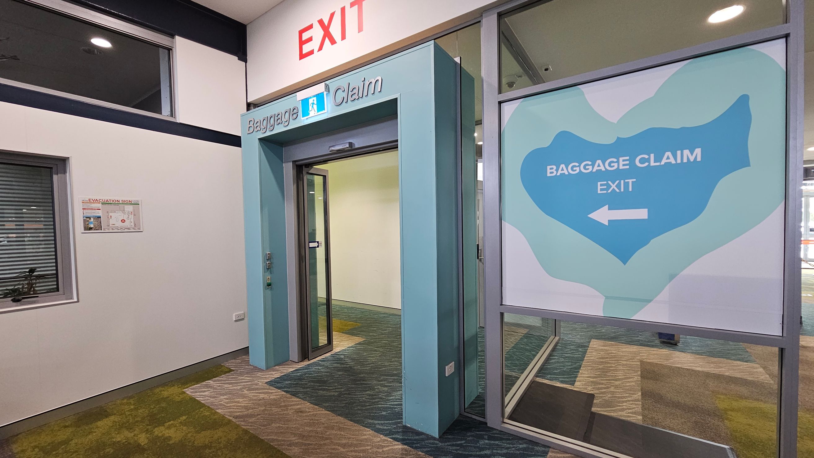 A corridor with a sign above reading "EXIT" in red. Below, a turquoise-framed doorway marked "Baggage Claim" leads to another area. To the right, a large sign with a blue shape and white text says "BAGGAGE CLAIM EXIT" with a left-pointing arrow. The floor is covered in a patterned carpet with green and grey tones. On the left wall, there's a window and an evacuation sign. The ceiling is light-coloured with recessed lighting.