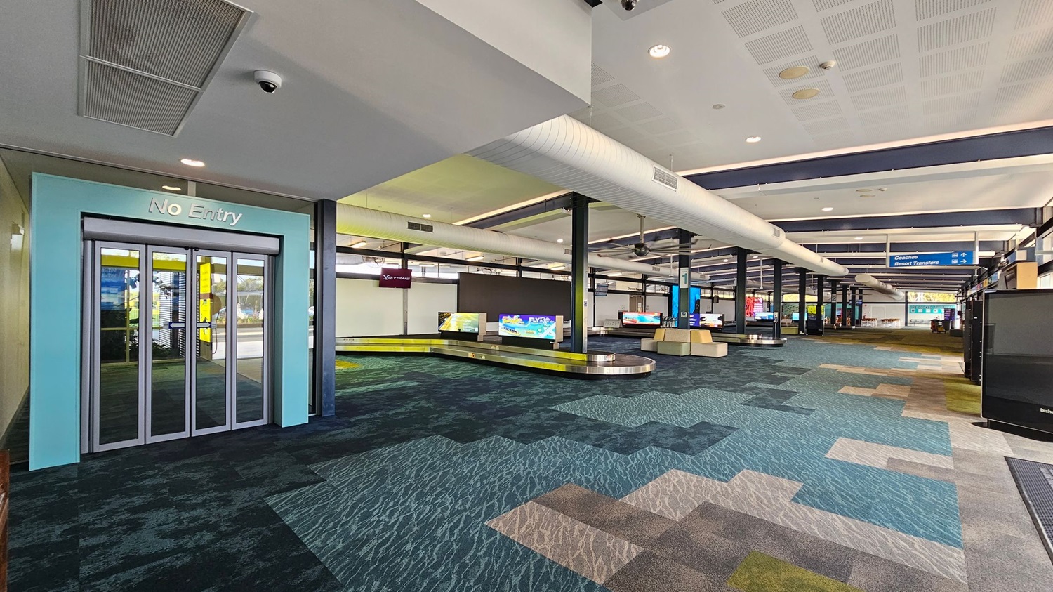 A spacious airport baggage claim area with a patterned carpet in shades of blue, green, and beige. On the left, there's a "No Entry" sign above a set of glass doors. In the centre, several baggage carousels are visible, each with digital screens displaying information. The ceiling features large white air ducts and recessed lighting. Towards the back, signs indicate directions for coaches and resort transfers. The area is well-lit and open.