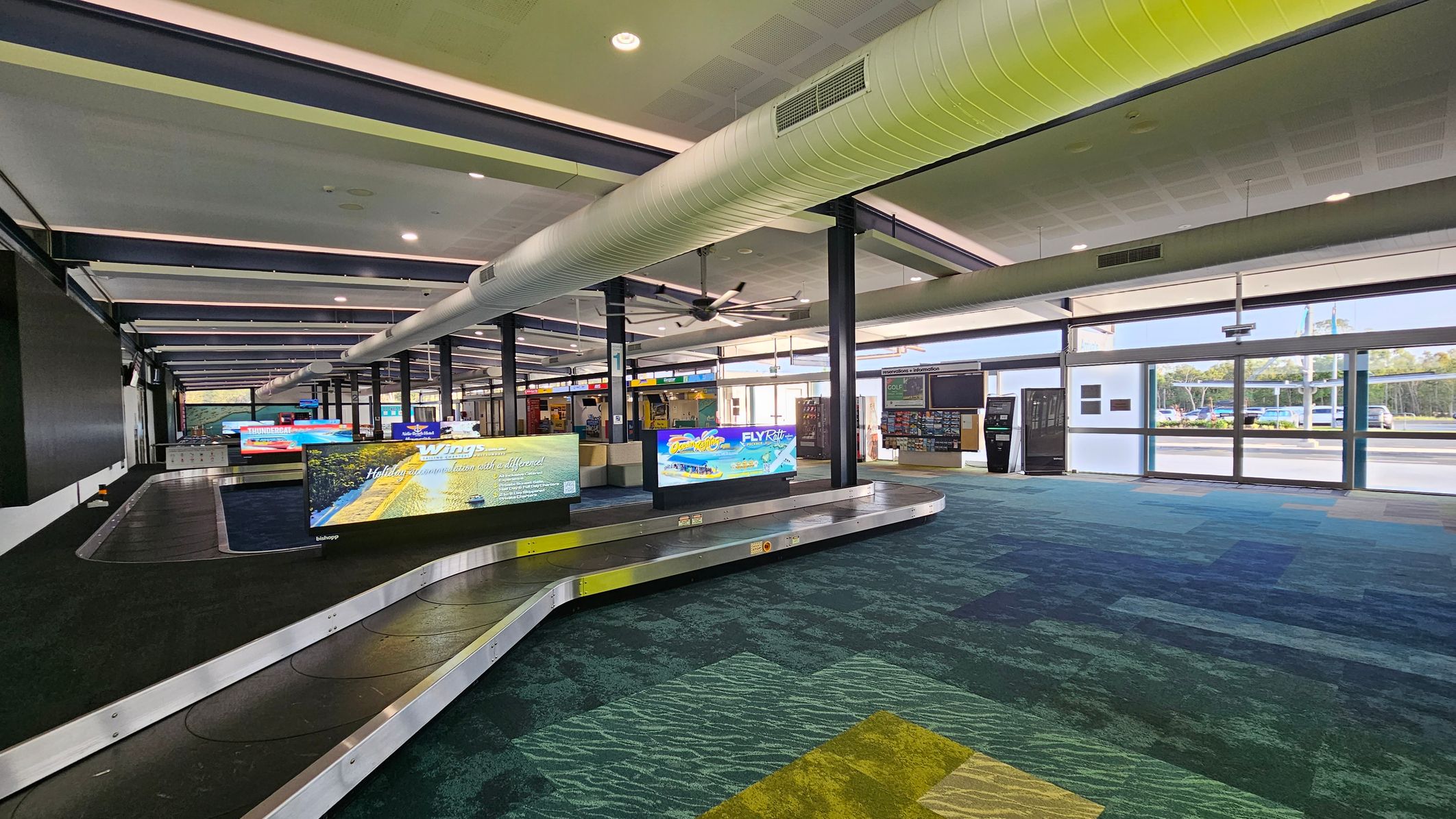 Interior of an airport baggage claim area. On the left, a conveyor belt with digital screens displaying advertisements. The floor is covered with a patterned carpet in shades of blue and green. The ceiling features exposed ductwork and lighting, with a large ceiling fan. On the right, large windows allow natural light in, with views of parked cars and trees outside. Vending machines and information boards are visible near the windows.