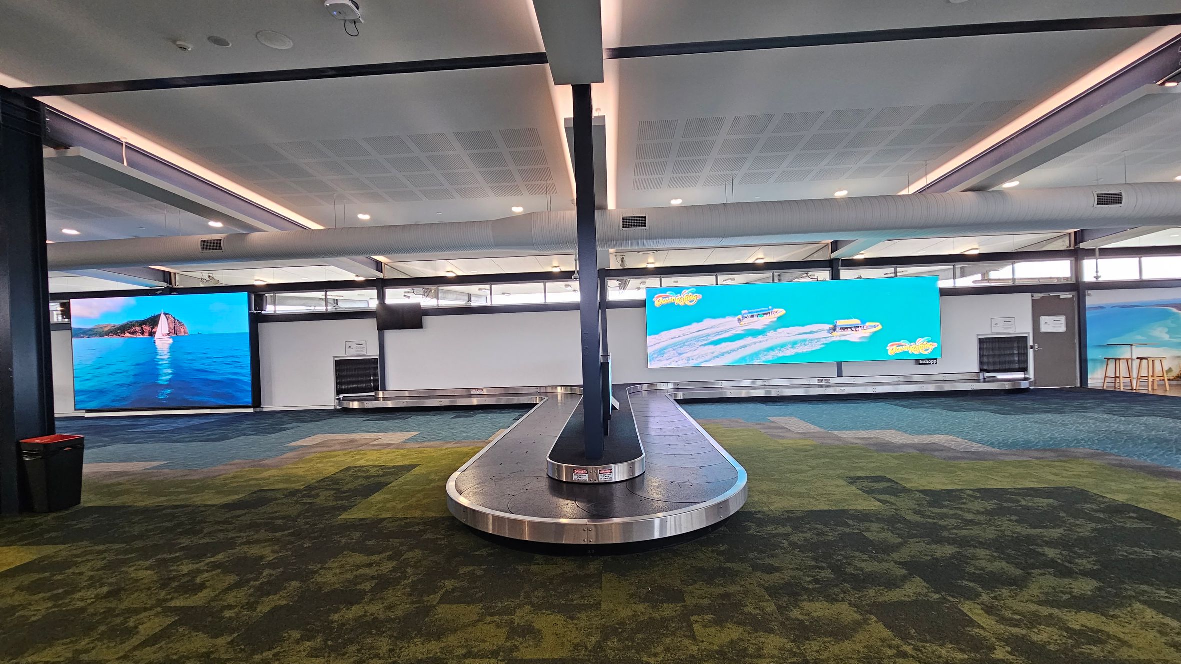 A baggage claim area with a conveyor belt in the centre. The floor is covered with a patterned carpet in shades of green and blue. On the left, a large screen displays an image of a sailboat on the sea. On the right, another screen shows speedboats on water with text. The ceiling is white with recessed lighting and visible air ducts. In the background, there are windows and a door on the right side.