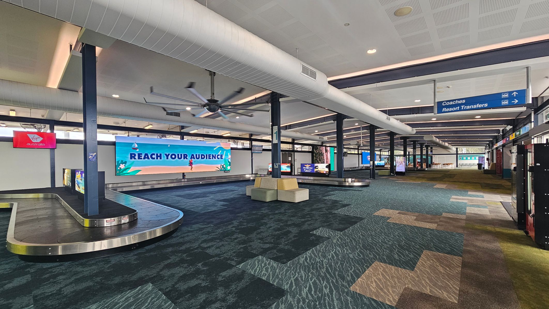 A spacious airport baggage claim area with a patterned carpet in shades of green, blue, and beige. On the left, there are luggage carousels with a large advertisement reading "REACH YOUR AUDIENCE." Overhead, a large ceiling fan and ductwork are visible. To the right, signs direct passengers to "Coaches" and "Resort Transfers." The ceiling is white with recessed lighting, and there are seating areas with cushioned benches near the carousels.