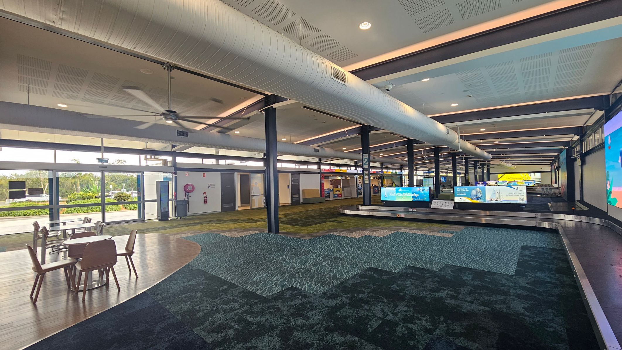 A spacious airport terminal with a high ceiling featuring large ventilation ducts and ceiling fans. On the left, there are tables and chairs on a wooden floor section. The right side has a baggage carousel with digital screens displaying advertisements. The floor is covered with a patterned carpet in shades of green and blue. Large windows on the left let in natural light, and there are various signs and facilities in the background.