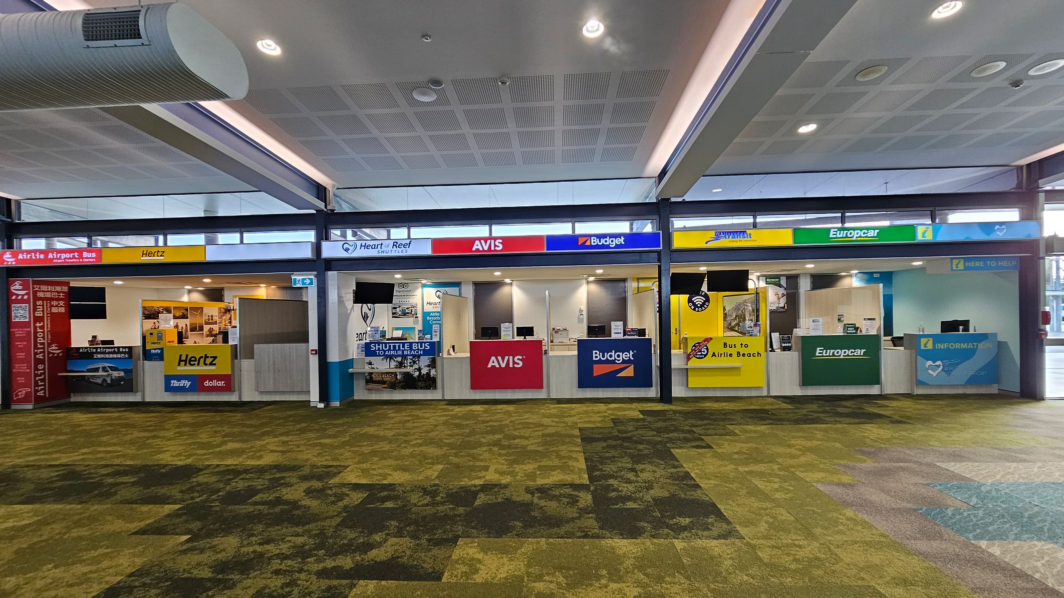 Car rental counters at an airport. From left to right: Hertz with a yellow sign, Avis with a red sign, Budget with a blue sign, and Europcar with a green sign. Above the counters are signs for shuttle services. The floor is covered in a patterned green and brown carpet. The ceiling is white with recessed lighting and a visible air duct.