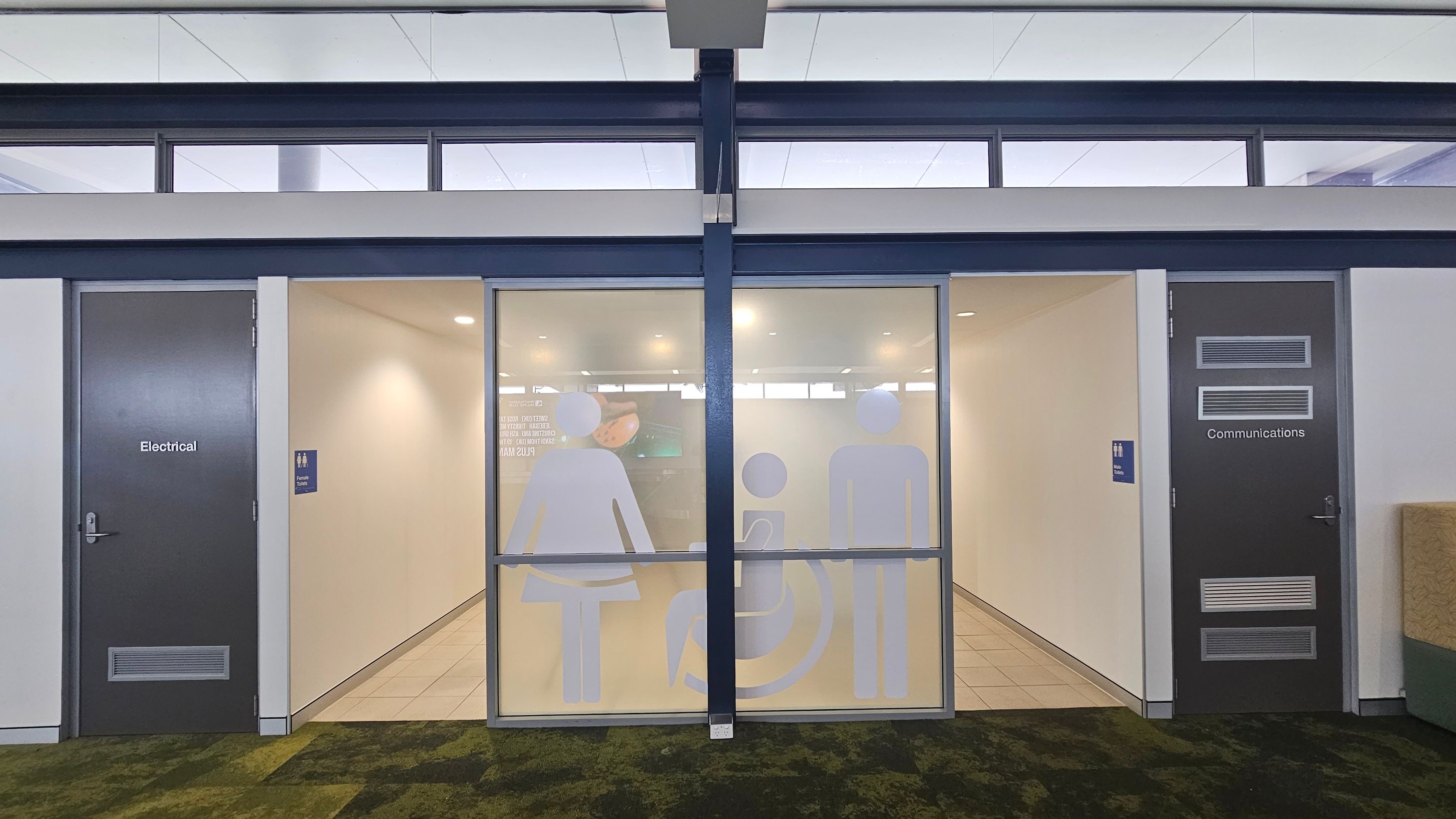 A hallway with three doors. The left door is labelled "Electrical" and the right door is labelled "Communications". The central area features frosted glass with large icons representing a woman, a person in a wheelchair, and a man. The floor is covered with a patterned carpet, and the ceiling has recessed lighting.