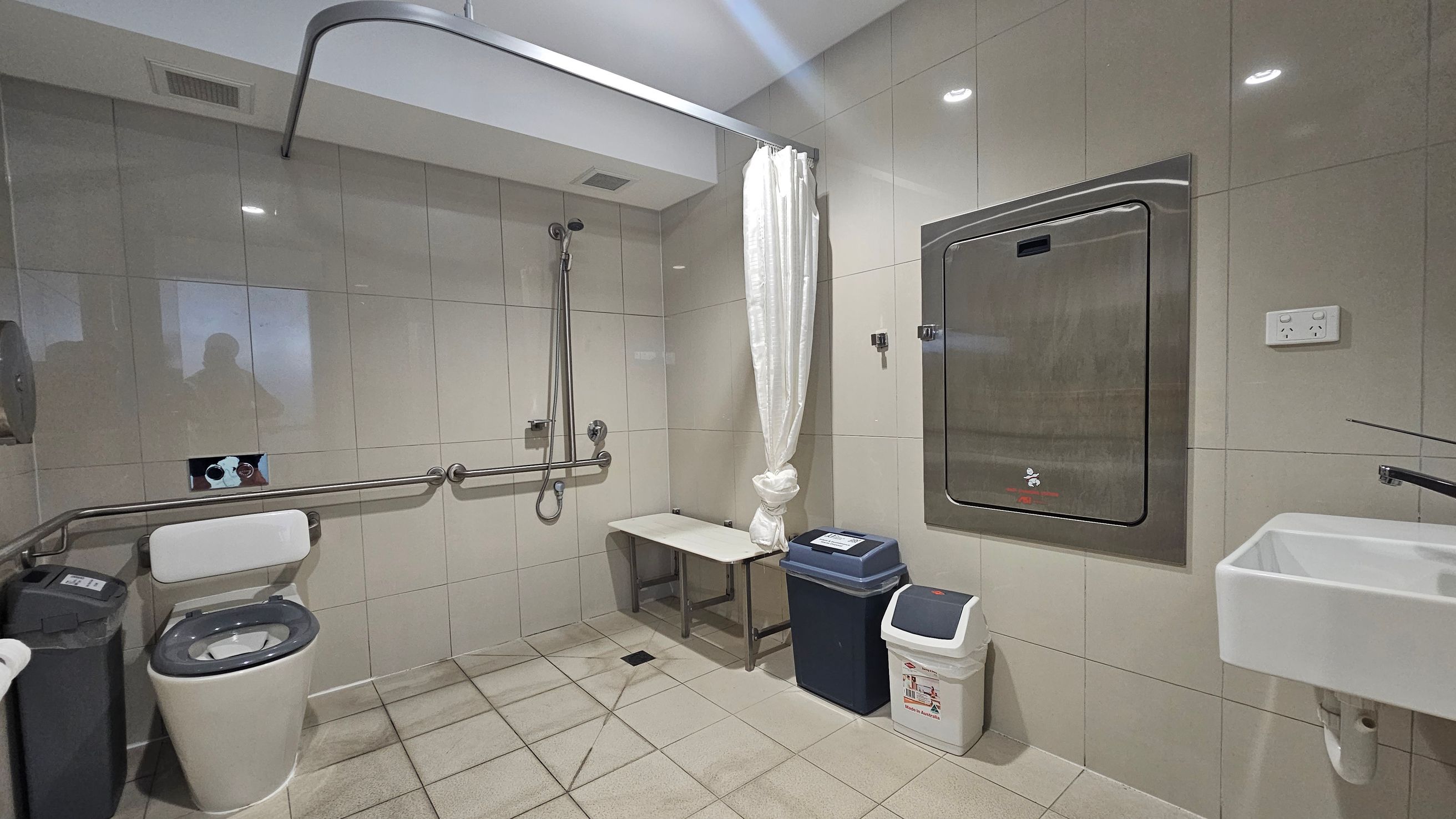 A spacious, accessible bathroom with light green tiled walls and a tiled floor. On the left, there is a toilet with a support rail and a bin nearby. In the centre, a shower area with a handheld showerhead and a bench is visible, separated by a white curtain. On the right, there is a stainless steel baby changing station mounted on the wall, a small sink, and two bins below. The ceiling is white with recessed lighting.