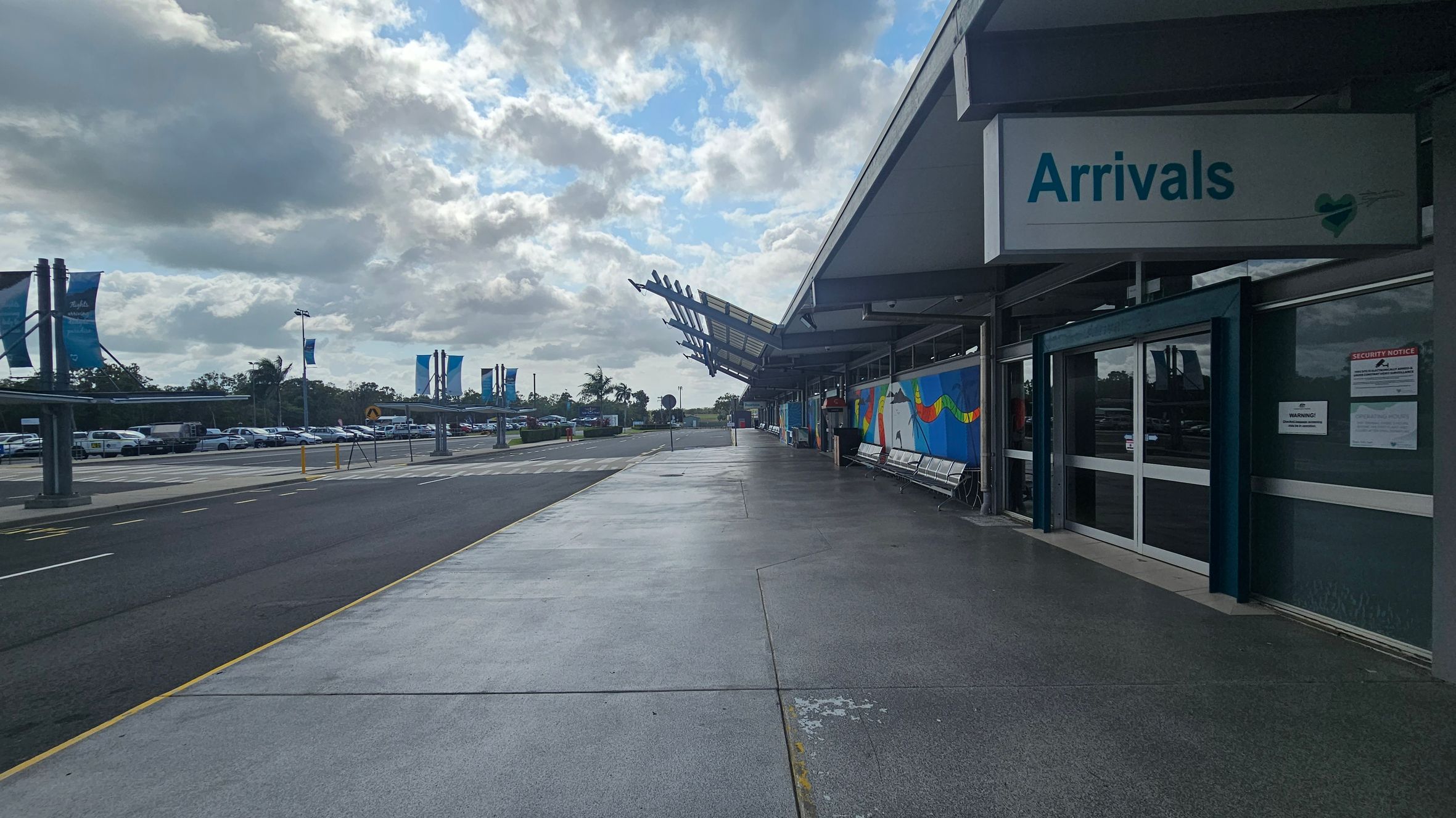 A concrete walkway leads to an airport arrivals area on the right, with a sign reading "Arrivals" above the entrance. The building features a modern design with a slanted roof and colourful murals on the walls. On the left, a car park is visible with rows of parked cars and tall banners. The area is open and spacious, with a clear path for pedestrians.