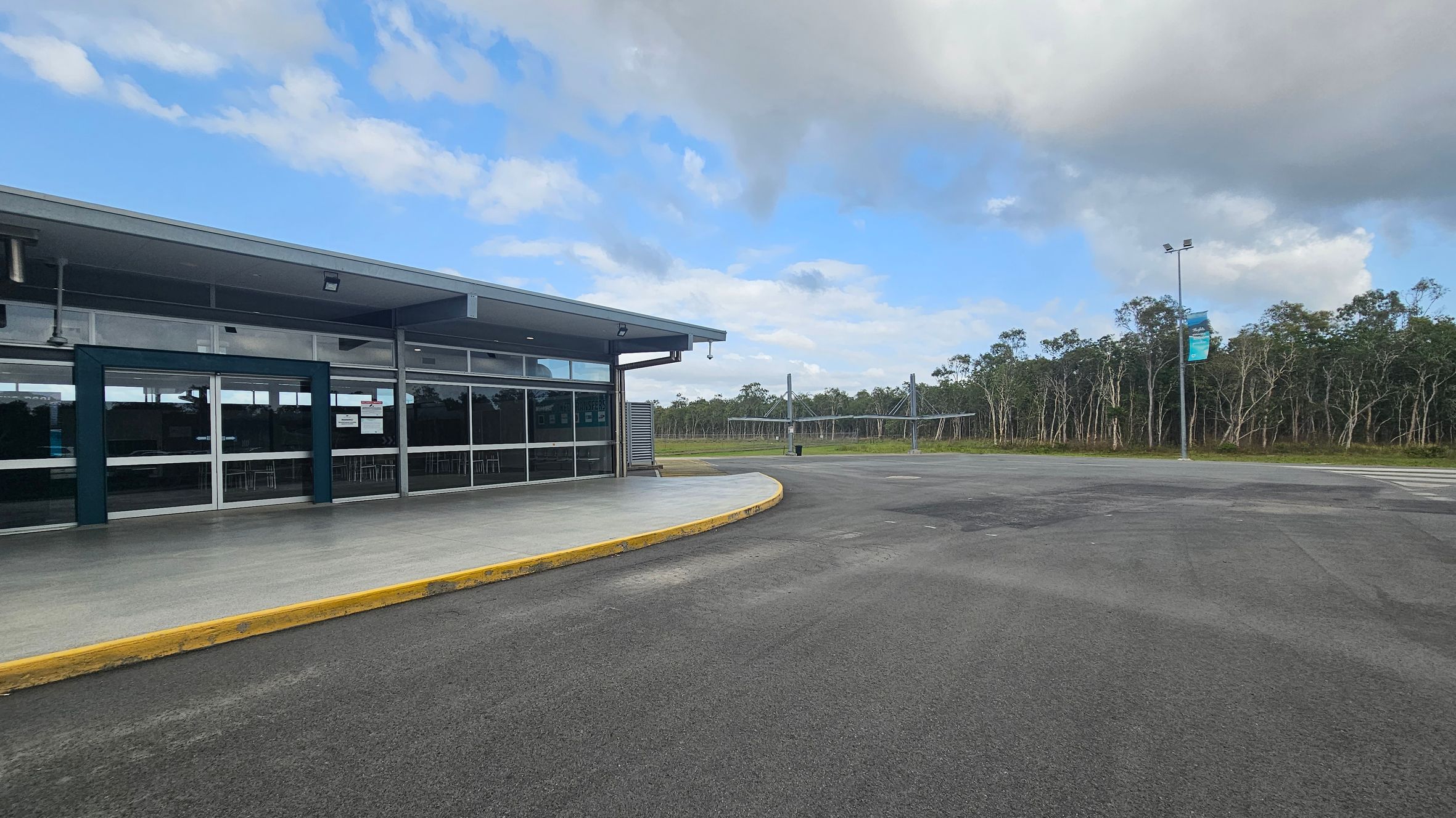 A modern building with large glass windows is on the left, featuring a covered walkway and a yellow kerb. The ground is paved with asphalt. In the background, there is a line of trees and a lamppost with a banner. The sky is partly cloudy.