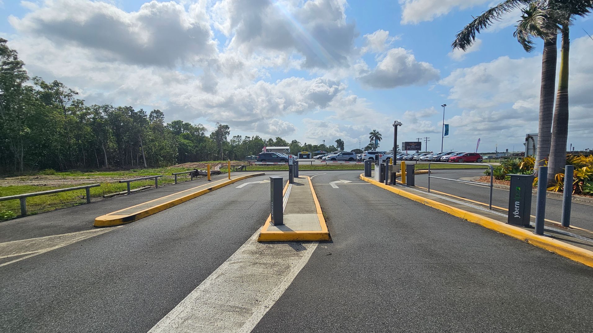 A parking lot entrance with two lanes separated by a yellow kerb. On the left, there is a grassy area with trees and a wooden fence. On the right, a palm tree and some shrubs are visible. In the background, several parked cars and a few signs can be seen. The road surface is asphalt, and there are directional arrows painted on the lanes.