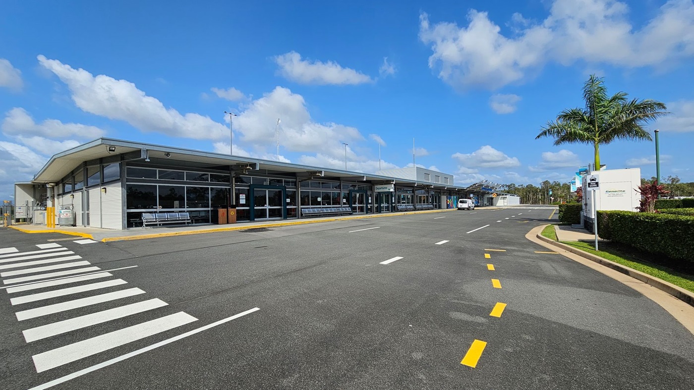 A modern, single-storey airport terminal with large glass windows and a flat roof is on the left. Several metal benches are visible outside. A zebra crossing leads towards the entrance. On the right, a palm tree stands next to a white sign and a hedge. The road in front has yellow and white markings, and a small white vehicle is parked further down. The background features a clear blue sky with scattered clouds.