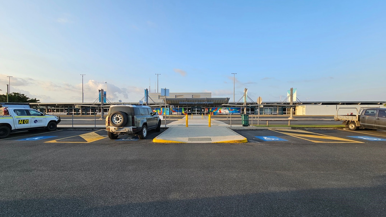 A parking area with two vehicles on either side, a white utility vehicle on the left and a grey SUV on the right. In the centre, a walkway with yellow bollards leads towards a building with a colourful mural. The building has a modern design with a flat roof and large windows. The ground is marked with parking lines and disabled parking symbols.
