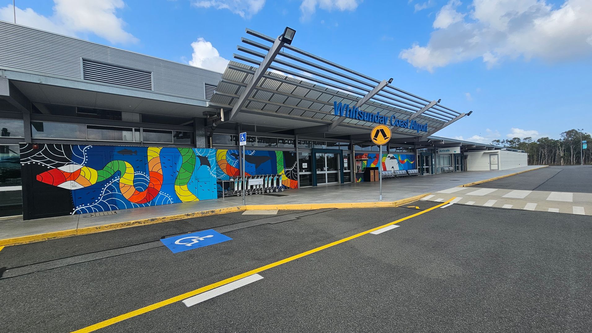 The image shows the entrance of Whitsunday Coast Airport. The building features a colourful mural with a snake design on the left. A row of luggage trolleys is positioned near the entrance. Above, a metal awning extends over the walkway. A pedestrian crossing sign is visible in front of the entrance. The ground has a marked parking space for disabled access, and the road has yellow and white lines. Trees are visible in the background on the right.