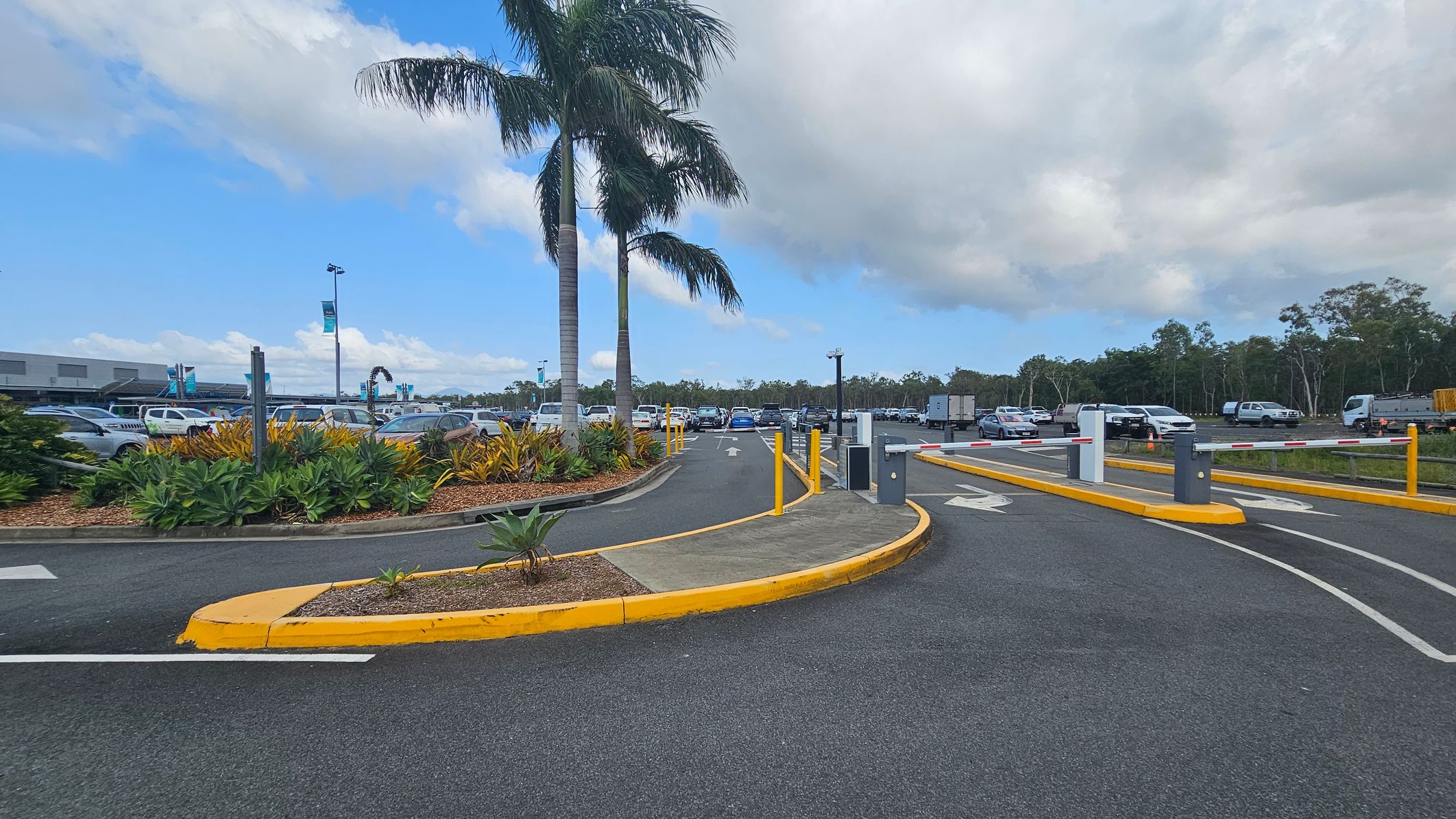 A car park entrance with two tall palm trees on the left, surrounded by green and yellow foliage. The road is divided by yellow kerbs, with arrows indicating directions. Several cars are parked in the background, and a few are driving towards the exit. The area is bordered by trees, and there are barriers at the entrance and exit points. The sky is partly cloudy.