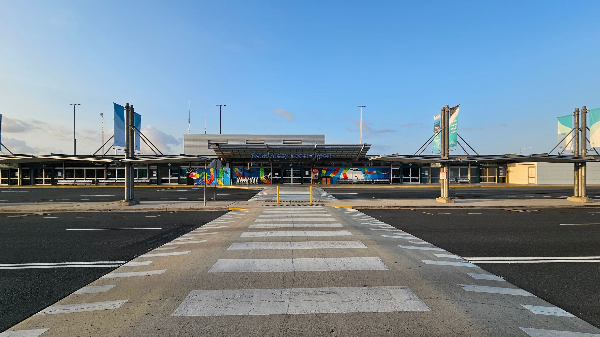 A wide view of an airport terminal with a modern design. In the centre, a pedestrian crossing leads to the entrance, which features a colourful mural. The terminal has a flat roof with a canopy extending over the entrance. On both sides, there are large signs supported by metal structures. The foreground shows a paved area with road markings, and the background includes a clear sky.