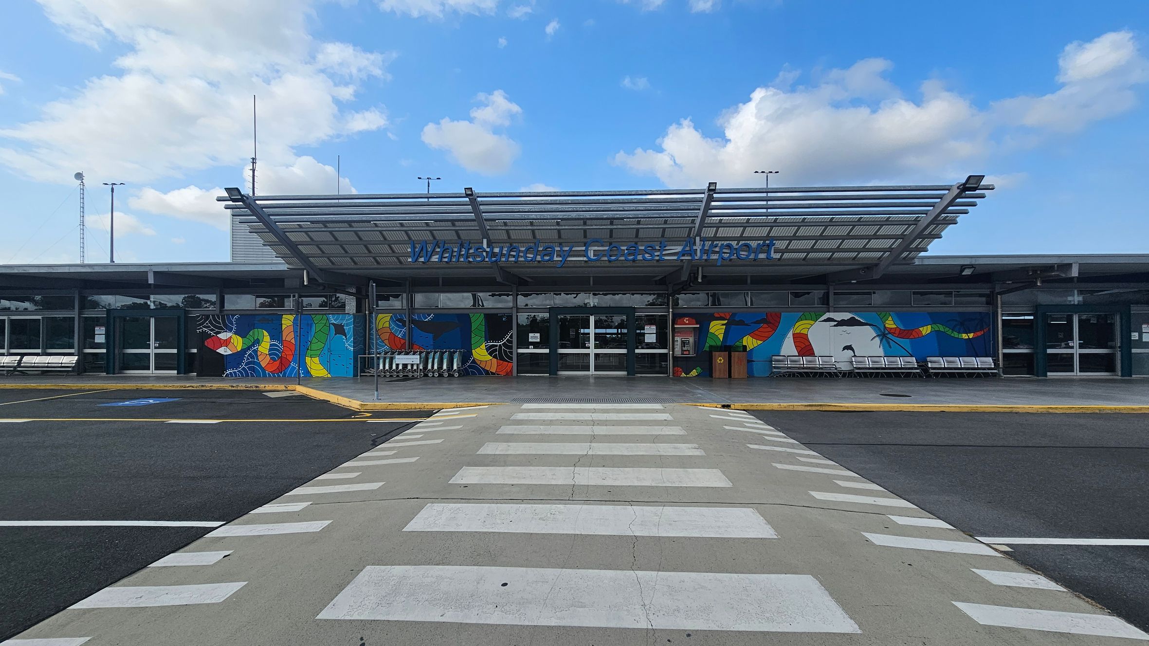 A modern airport entrance with a large glass facade and a metal canopy. The building features colourful murals with abstract designs on either side of the entrance. A row of trolleys is visible to the left, and a line of benches is on the right. The foreground shows a pedestrian crossing leading to the entrance. The ground is paved with asphalt and concrete.