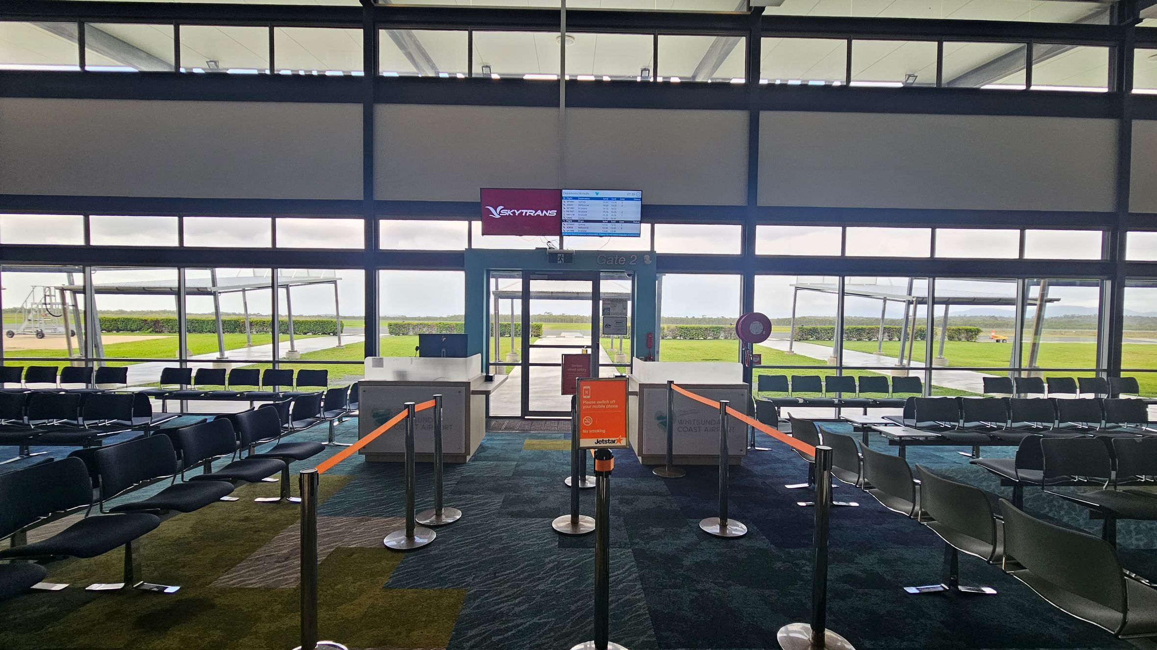 An airport gate area with rows of black seating on a patterned carpet. In the centre, there are barriers with orange tape leading to a boarding gate. Above the gate, a screen displays flight information with the Skytrans logo. Large windows in the background reveal a view of the tarmac and green grass outside. The ceiling is high with visible beams.