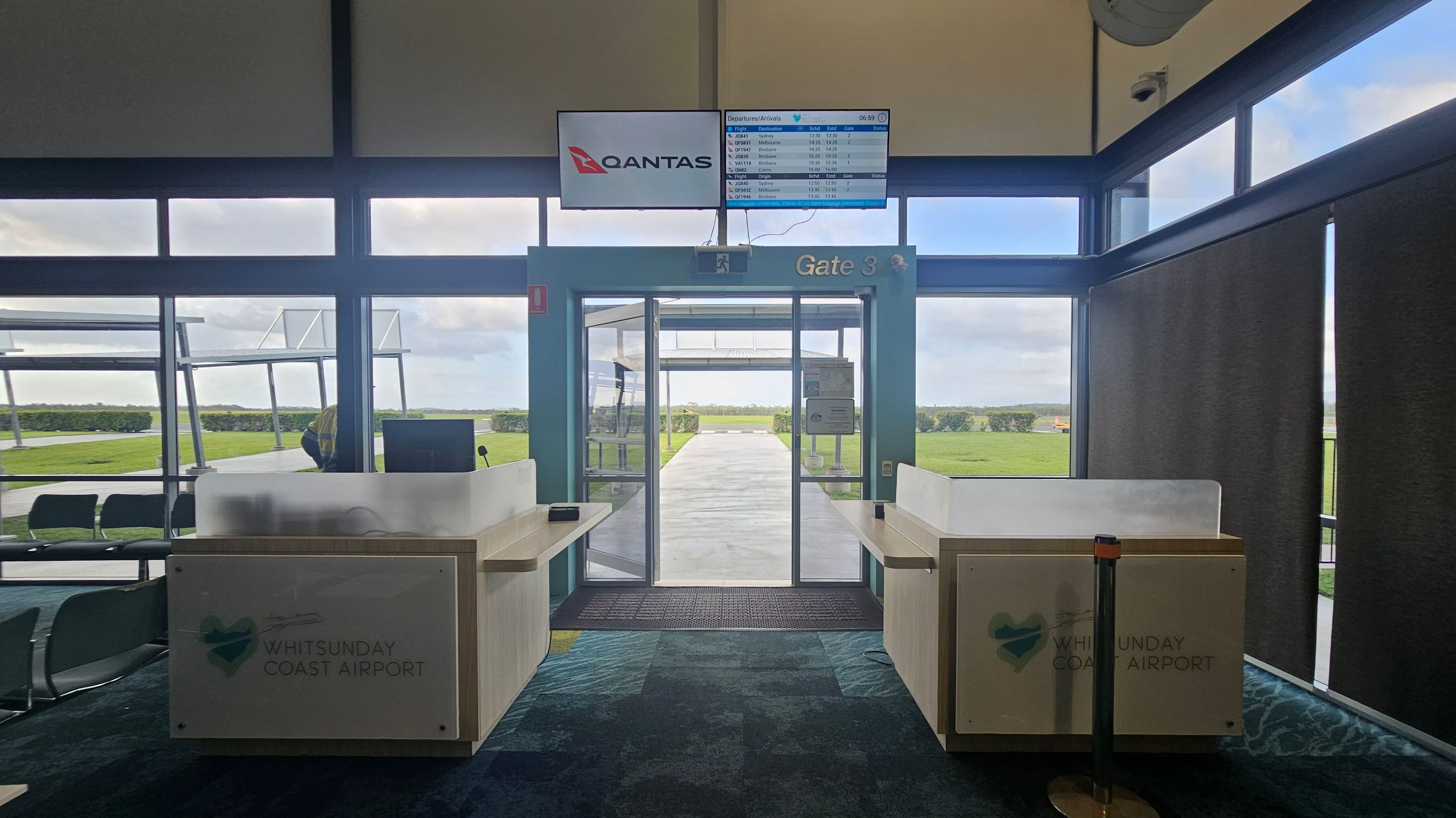 Inside an airport terminal at Gate 3, with a Qantas sign and a departures/arrivals screen above. Two check-in counters are on either side, marked with "Whitsunday Coast Airport". Large windows provide a view of the tarmac and grassy area outside. The floor is carpeted in a blue and green pattern, and the ceiling is high with exposed beams.