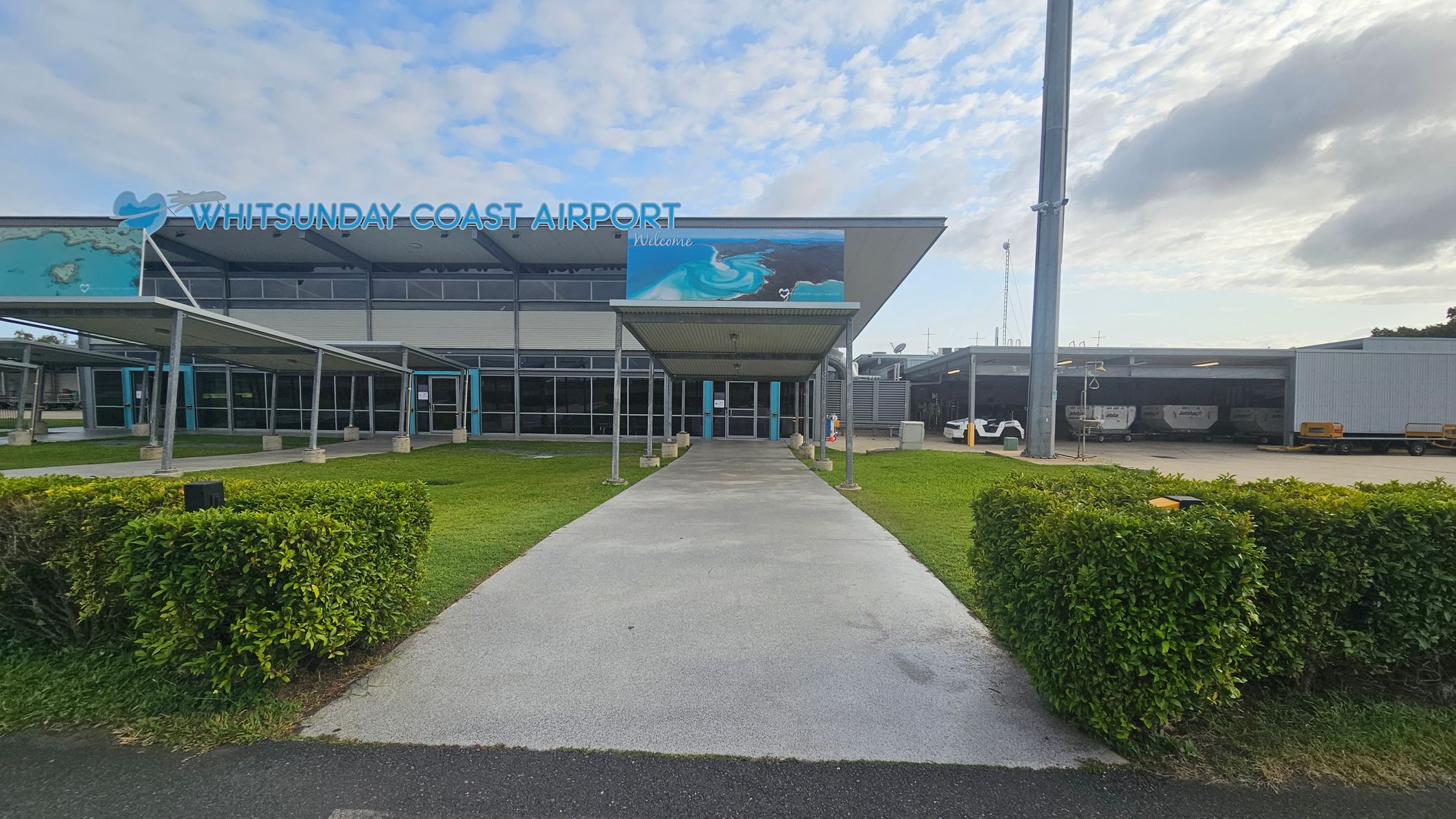 The image shows the entrance to Whitsunday Coast Airport. In the centre, a covered walkway leads to glass doors. The building has a large sign with the airport's name and a mural depicting ocean waves. On the left and right, there are green hedges and grass. To the right, a large pole and a covered area with vehicles are visible. The building has a modern design with a flat roof and large windows.