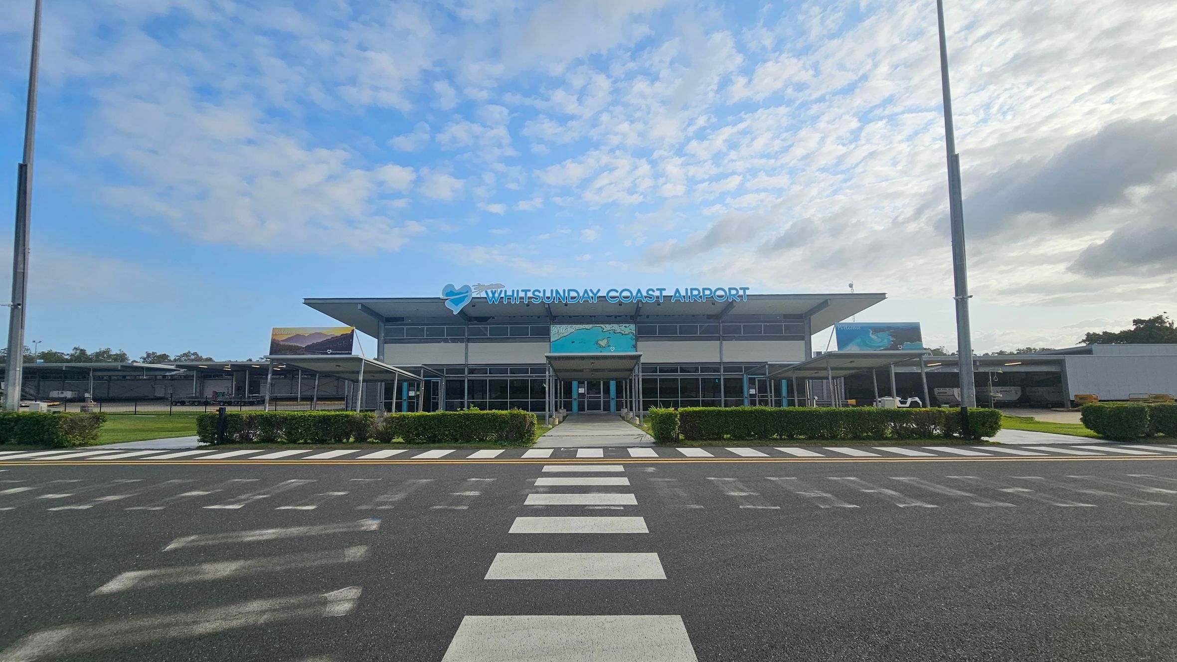 A modern airport terminal with a flat roof and large glass windows. The sign reads "Whitsunday Coast Airport" in blue letters. In front, there is a pedestrian crossing on an asphalt road leading to the entrance. Green hedges line the pathway, and there are covered walkways on either side. Two tall lampposts are visible, one on each side of the image. The background includes a few trees and a partly cloudy sky.