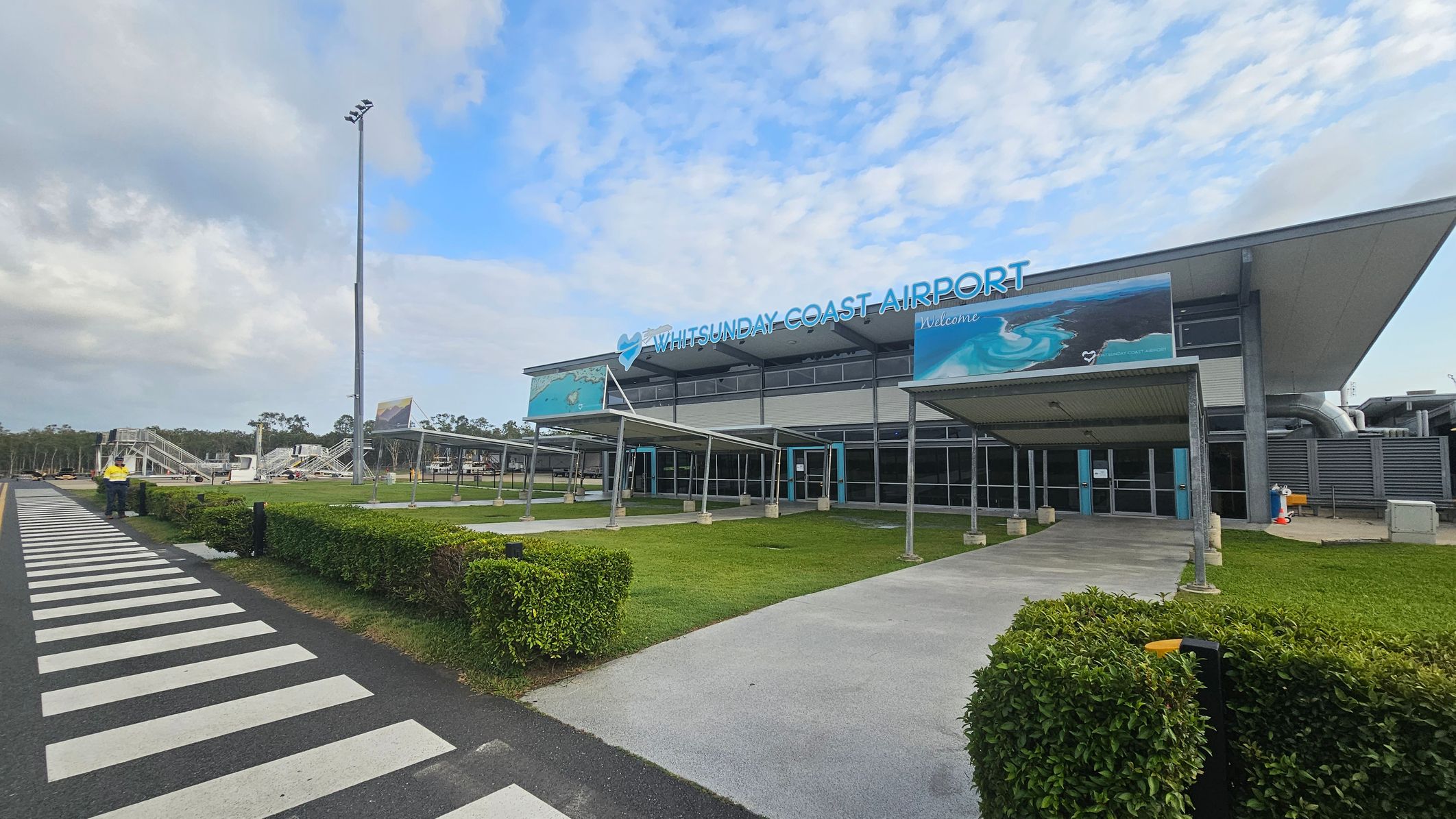 Whitsunday Coast Airport building with a large sign on the roof. The entrance features a covered walkway and glass doors. A zebra crossing leads to the building, bordered by neatly trimmed hedges. On the left, there are metal structures and a tall light pole. The ground is a mix of grass and paved paths.
