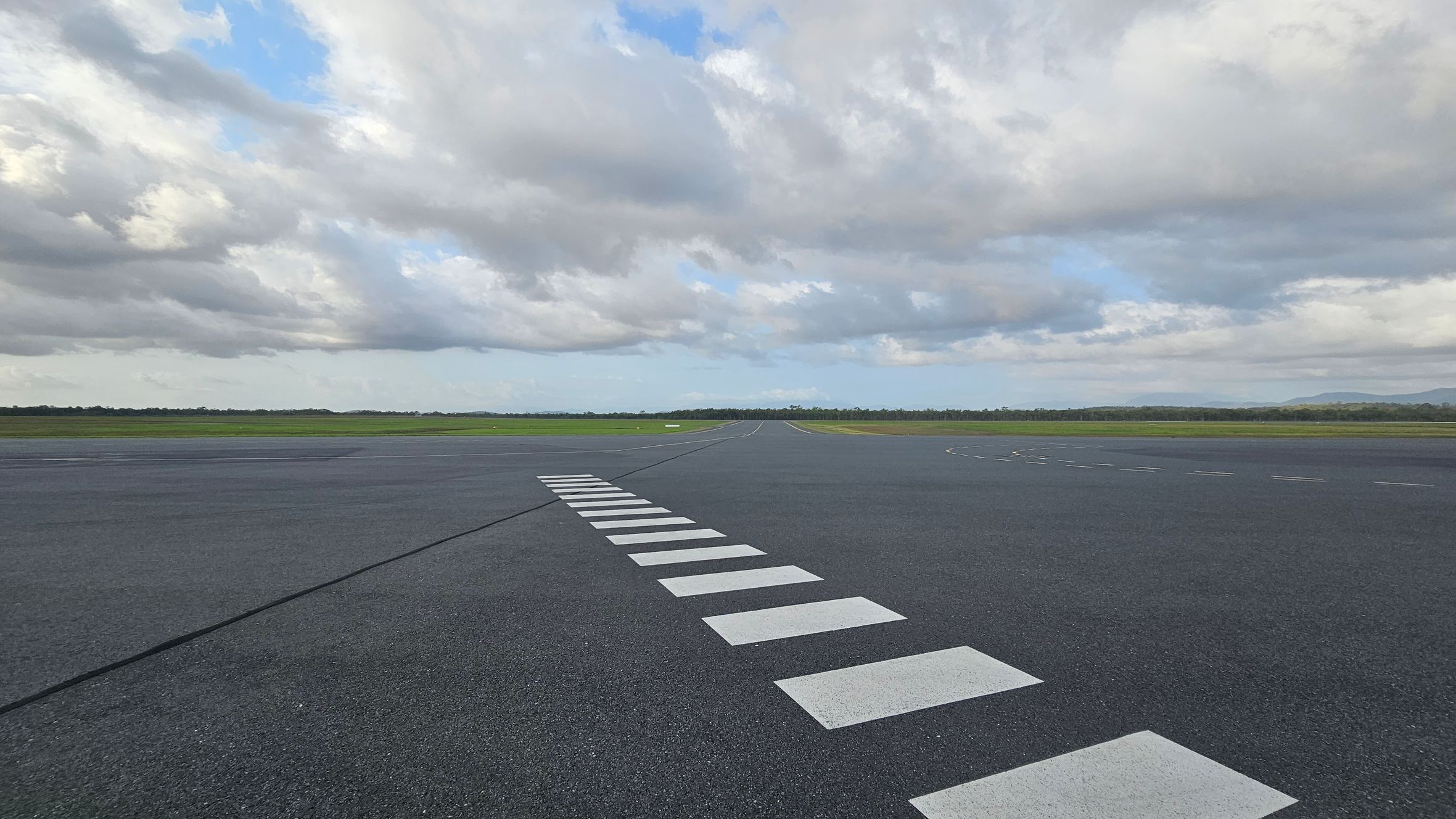A runway with a series of white rectangular markings leading into the distance. The surface is dark grey asphalt. In the background, there is a flat expanse of green grass and a line of trees. The sky is filled with large, fluffy clouds, with patches of blue visible.