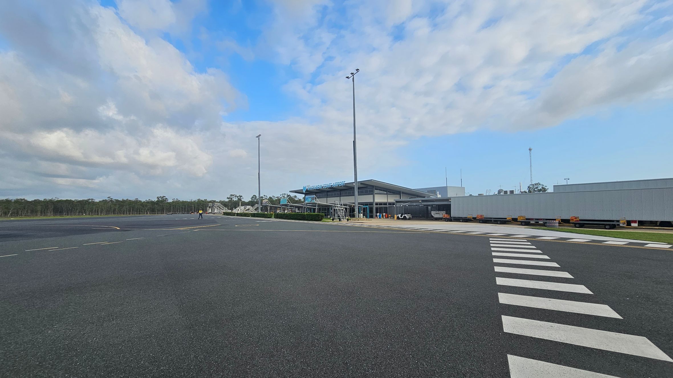 A wide view of an airport tarmac with a building labelled "Whitsunday Coast Airport" in the centre. The building has a modern design with a large overhanging roof. To the right, there are several trailers lined up. A pedestrian crossing is visible in the foreground, leading towards the building. On the left, a person in a yellow vest walks across the tarmac. The background features a line of trees under a partly cloudy sky.