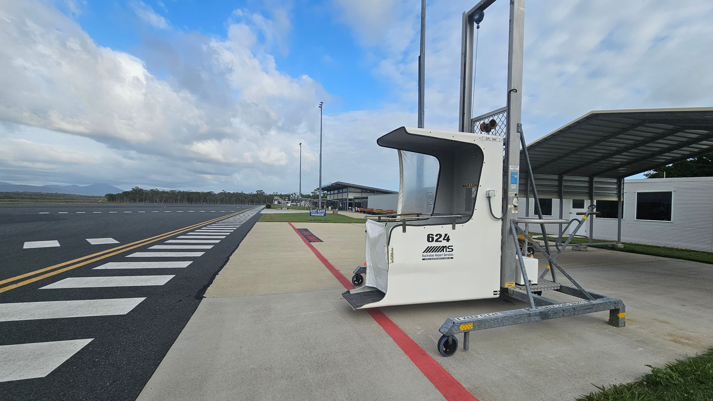 A small airport scene with a mobile passenger boarding ramp marked "624" on the right. The ramp is positioned on a concrete surface with a red line. In the background, there is a covered structure and a building with windows. On the left, a runway with white markings stretches into the distance, bordered by trees. The sky is partly cloudy.