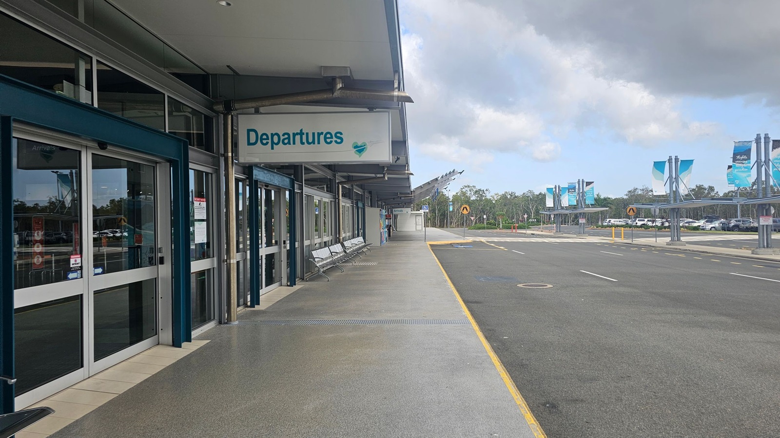 On the left, there are glass sliding doors with a sign above reading "Departures" in blue. Metal benches are placed along the wall. The floor is a mix of grey and green tiles. On the right, there is a wide road with a yellow line marking the edge. In the background, there are several blue and white banners on poles, and a parking area with cars. The ceiling is flat and grey.