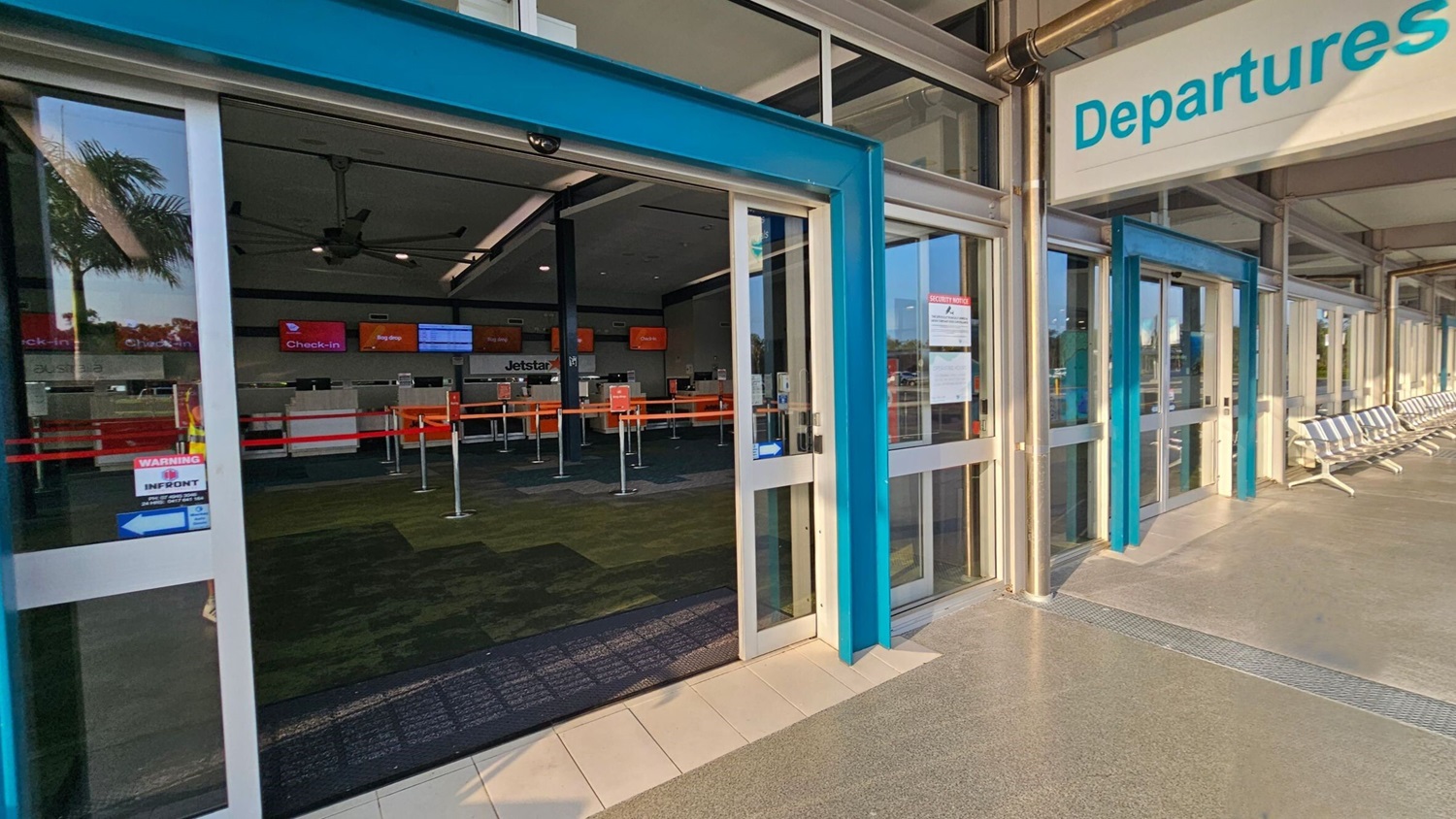 Automatic sliding glass doors lead into an airport departures area. Inside, check-in counters with red barriers and digital screens are visible. The floor is a mix of tiles and carpet. Outside, a row of metal benches lines the right side. A shadow of a person is cast on the ground in the foreground. A palm tree is reflected in the glass on the left. The ceiling inside has large fans.