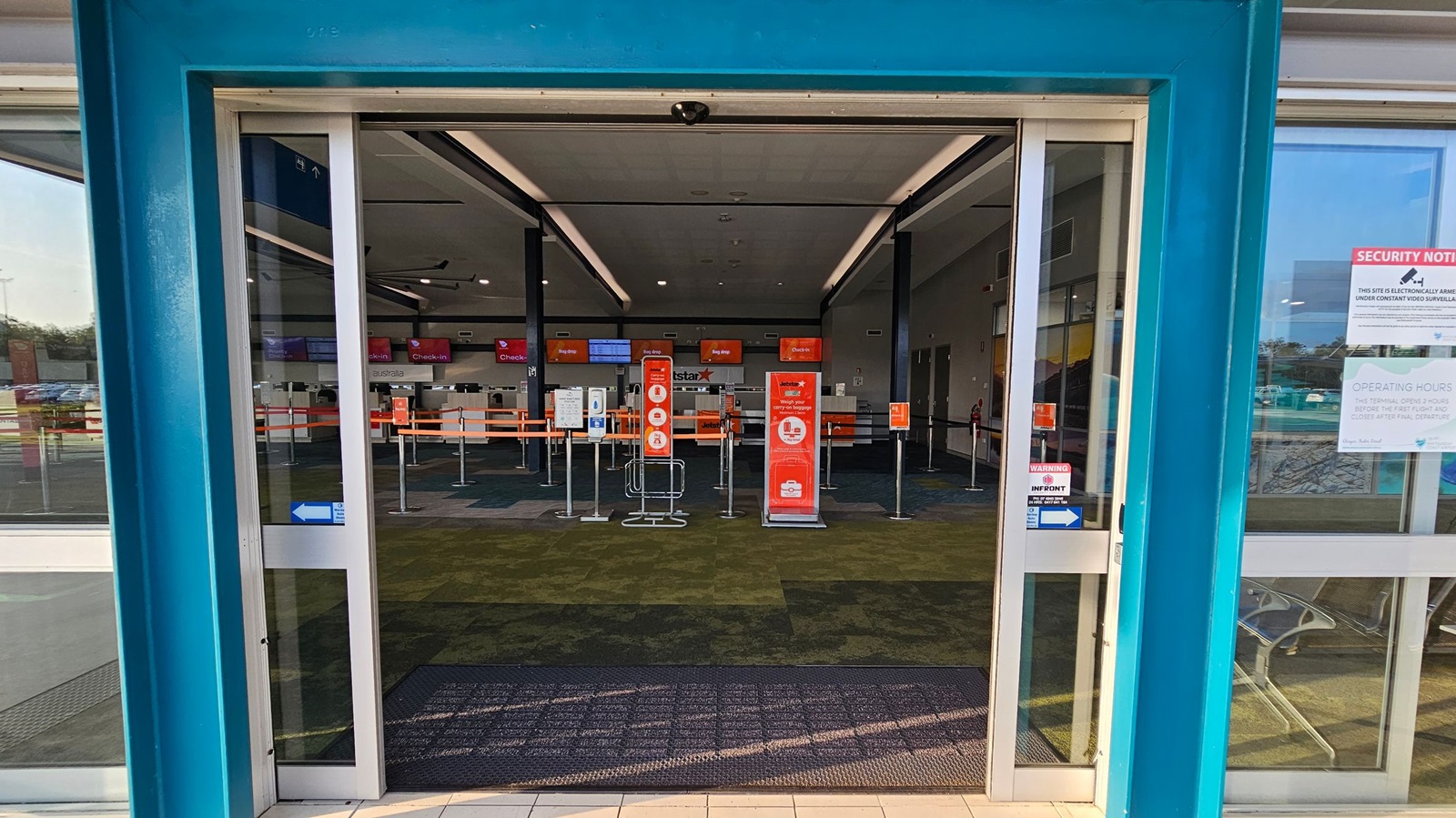Automatic sliding glass doors open to reveal an airport check-in area. Inside, there are multiple check-in counters with red signage and digital screens displaying "Check-in" and "Bag Drop". The floor is covered with a patterned carpet in shades of green and brown. Queue barriers guide passengers. On the right, a window displays security and operating hours notices. The ceiling has recessed lighting and black beams.