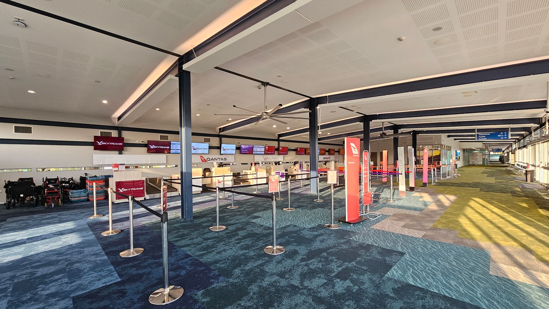 An airport check-in area with counters for Qantas and Skytrans on the left. The counters have digital screens above them displaying flight information. There are retractable barriers forming queues in front of the counters. The floor is covered with a patterned carpet in shades of blue and green. The ceiling is high with exposed beams and a large ceiling fan. On the right, there are windows allowing natural light to illuminate the space, and signs indicating directions to departure gates.