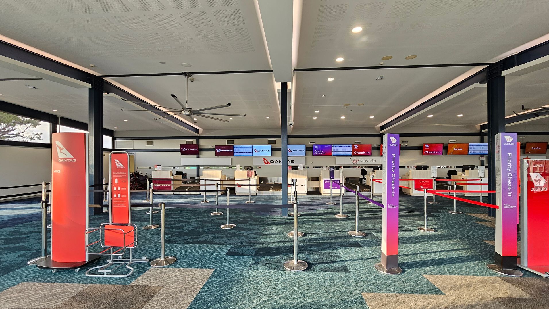 An airport check-in area with multiple counters in the background, featuring Qantas and Virgin Australia signage. The floor has a patterned carpet in shades of blue and beige. In the foreground, there are red and purple signs for Qantas and Virgin Australia, indicating service and priority check-in areas. The ceiling is light-coloured with recessed lighting and a large ceiling fan. Queue barriers are set up with retractable belts.