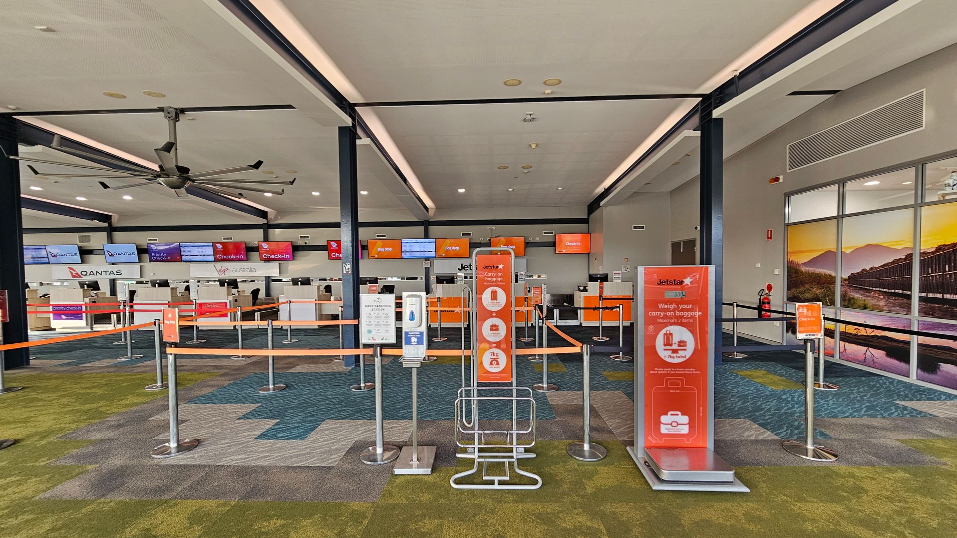 An airport check-in area with multiple counters in the background, including Qantas and Jetstar. Digital screens display check-in information above the counters. In the foreground, there are signs and barriers for queue management, including a Jetstar sign about carry-on baggage. The floor is carpeted in a mix of green and blue patterns. The ceiling is white with recessed lighting and a large ceiling fan. A mural of a scenic landscape is visible on the right wall.