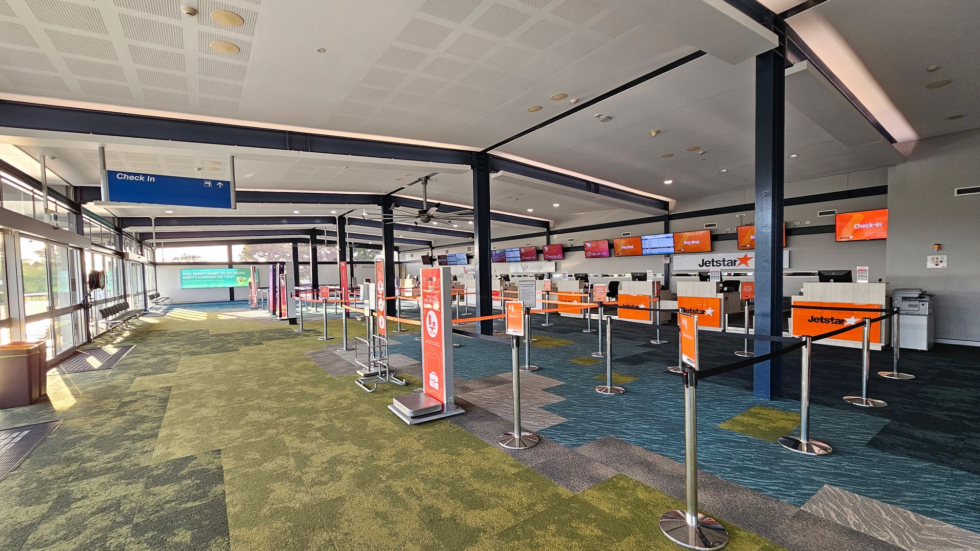An airport check-in area with multiple counters on the right, each displaying orange signs with "Jetstar" and "Check-in". The counters are separated by retractable barriers. On the left, there are large windows allowing natural light in, with a row of seats and a blue "Check In" sign hanging from the ceiling. The floor is covered with a patterned carpet in shades of green and blue. The ceiling is white with recessed lighting.
