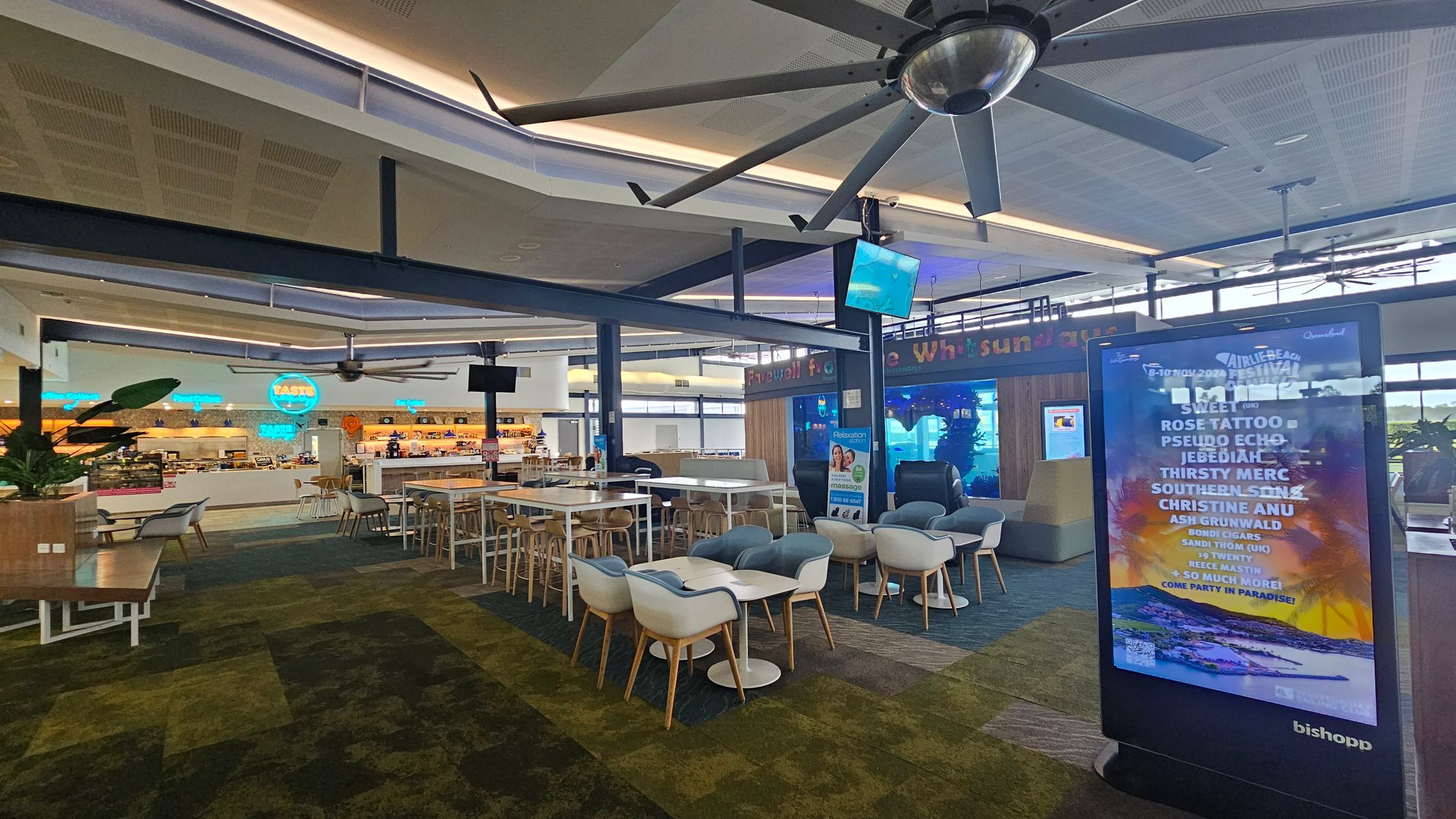 A spacious indoor area with a large ceiling fan and modern seating arrangements. On the right, there's a digital display board with event information. In the centre, there are tables and chairs, including high stools and cushioned seats. The left side features a food court with neon signs and a variety of food stalls. The floor is carpeted in a mix of green and brown tones, and the ceiling has a grid pattern with recessed lighting.