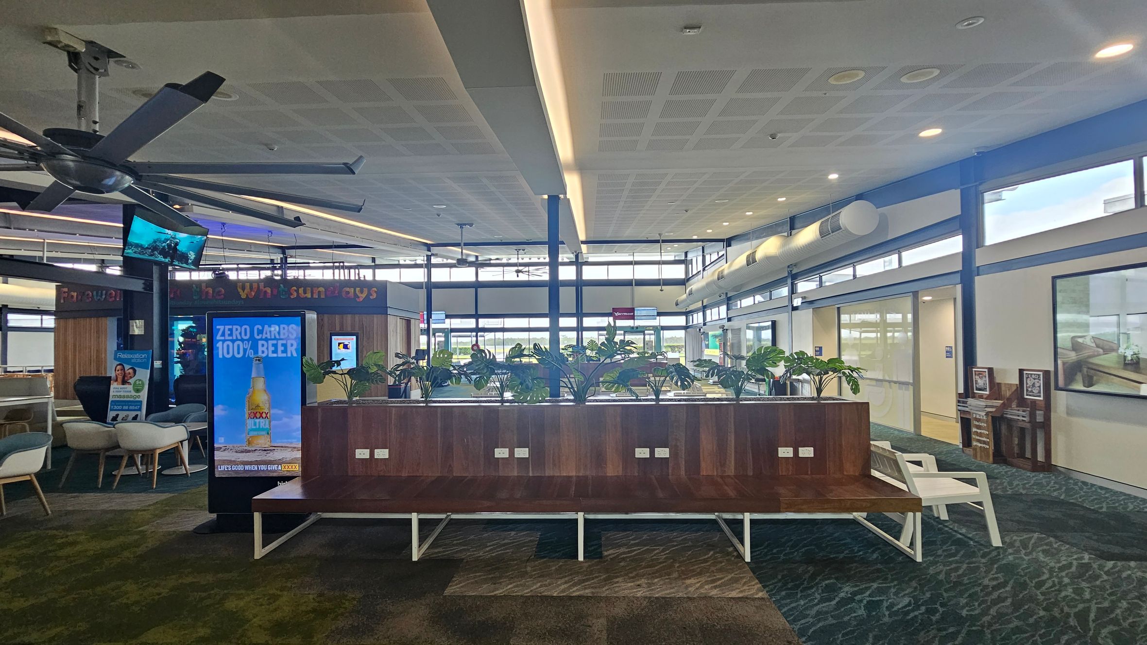 Interior of an airport lounge with a large ceiling fan on the left. A digital advertisement for beer is displayed on a screen in the centre. Behind it, a wooden partition with potted plants on top. The floor is carpeted in a green and grey pattern. On the right, there are white chairs and a wall with framed pictures. The ceiling has recessed lighting and a grid pattern. Large windows in the background allow natural light to enter.
