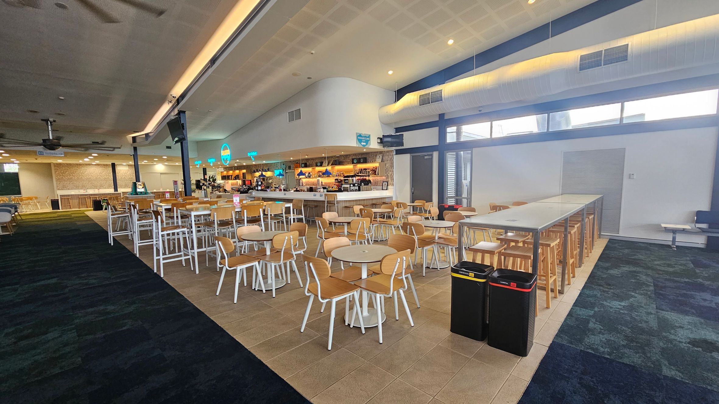 A spacious indoor dining area with a variety of seating options. On the left, there are round tables with wooden chairs and a long bar counter in the background. The right side features a long high table with stools. The floor is a mix of tiled and carpeted sections, and the ceiling is high with recessed lighting. Two bins are placed near the high table. The area is well-lit with natural light coming through windows on the right.