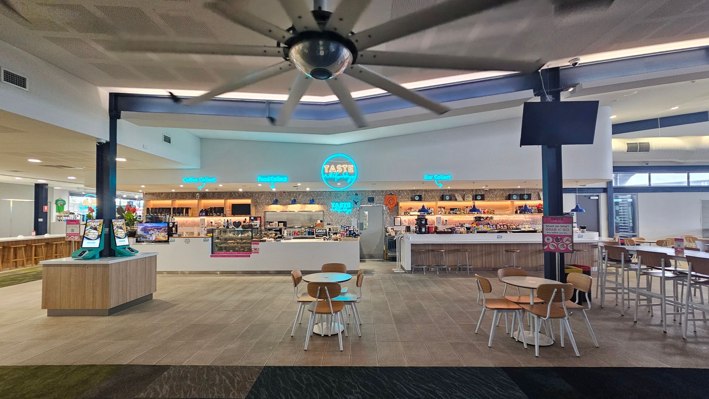 A spacious indoor café area with a large ceiling fan. In the centre, there's a counter with a neon sign reading "TASTE" and various food and drink options displayed. To the left, there are self-service kiosks on a wooden stand. On the right, a bar area with high stools and tables. The floor is tiled, and there are several round tables with wooden chairs scattered around. The ceiling has recessed lighting, and the overall atmosphere is modern and inviting.