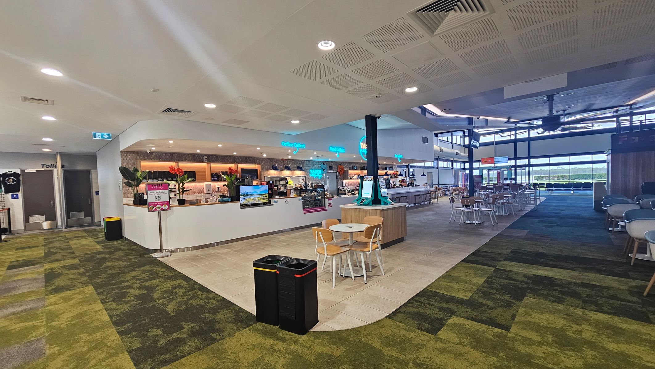A spacious indoor area with a café counter in the centre, featuring a variety of drinks and snacks. To the left, there are signs for toilets and a green-patterned carpet. In the foreground, two black bins and a small table with chairs are visible. The right side has more seating with tables and chairs, and large windows in the background allow natural light to enter. The ceiling is fitted with lights and ventilation panels.