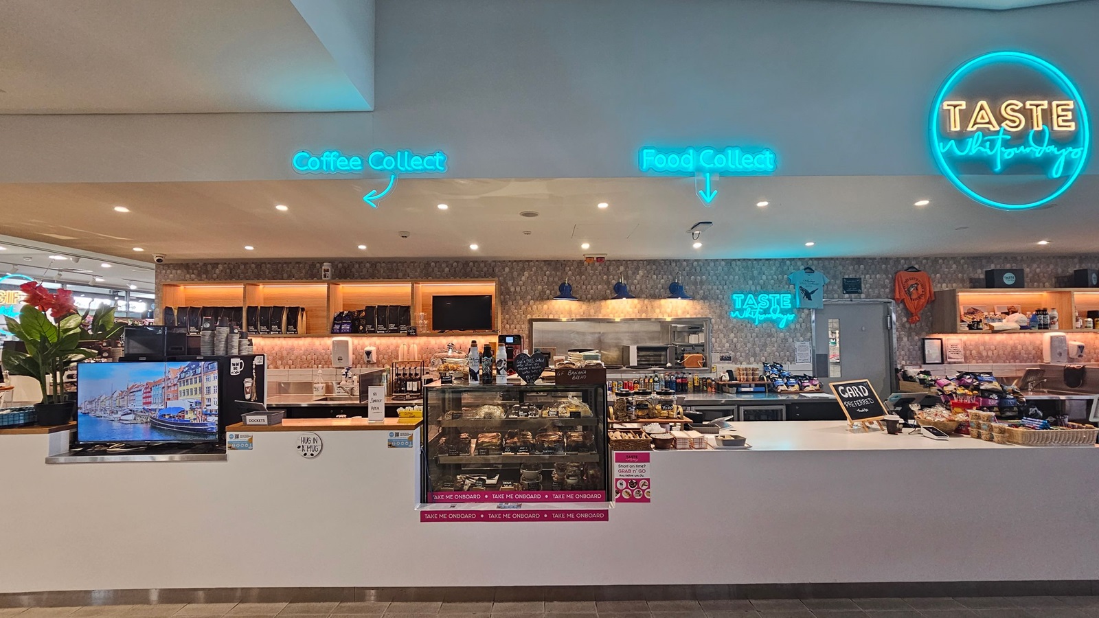 A modern café counter with a white surface. On the left, a TV screen displays a colourful cityscape. Above, neon signs read "Coffee Collect" and "Food Collect" with arrows. The middle section has a glass display filled with pastries and snacks, labelled "Take Me Onboard." Behind, shelves hold coffee bags and other items. On the right, a neon sign says "Taste" with a slogan. The ceiling is white with recessed lighting, and the floor is tiled.