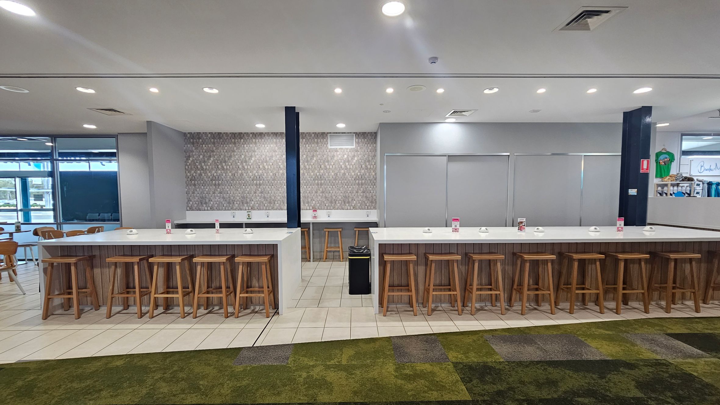 A modern indoor seating area with two long white tables, each lined with wooden stools. The tables have small white bowls and menu holders. The background features a grey textured wall and a section with closed blinds. The floor is a mix of tiled and green carpeted areas. Ceiling lights are evenly spaced above, and a window with a view of the outside is on the left. A small shop with clothing is visible on the right.