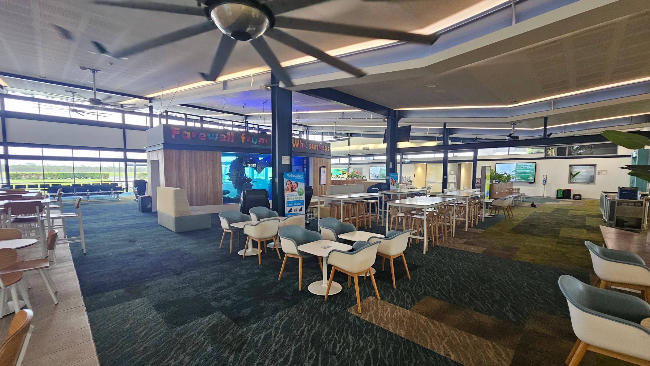 A spacious airport lounge with a mix of seating options, including round tables with chairs and long tables with stools. The floor is covered in a patterned carpet with shades of green and blue. A large ceiling fan is visible above. In the background, there are large windows allowing natural light in, and a wooden structure with a sign that reads "Farewell from Whitsundays." The ceiling has modern lighting fixtures, and there are plants and digital screens around the area.