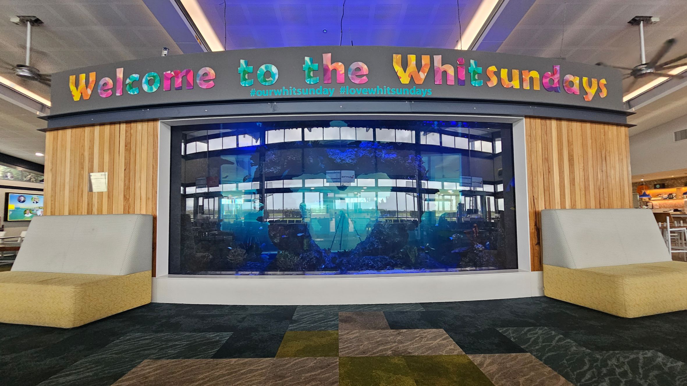A large aquarium is set into a wooden panelled wall with colourful letters above spelling "Welcome to the Whitsundays" and hashtags below. On either side of the aquarium are cushioned seats with light green backs and beige bases. The floor is carpeted in a patchwork of dark and light grey patterns. The ceiling is grey with recessed lighting and ceiling fans. In the background, there are tables and chairs, suggesting a café or waiting area.