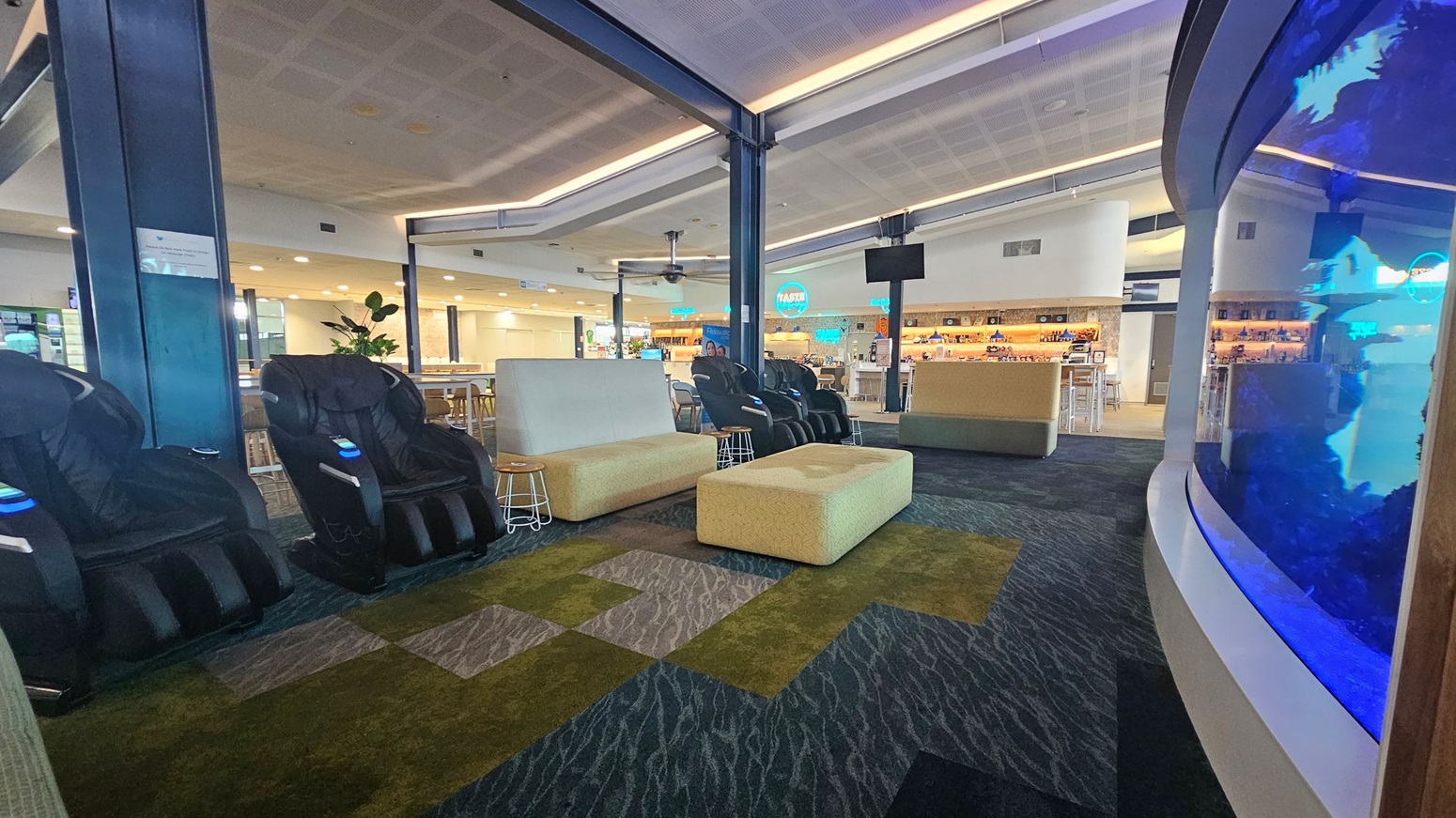 A modern lounge area with massage chairs on the left, upholstered seating in the centre, and a large curved aquarium on the right. The floor is covered with a patterned carpet in shades of green and grey. In the background, there is a bar with stools and a variety of drinks on display. The ceiling is fitted with recessed lighting and a ceiling fan.