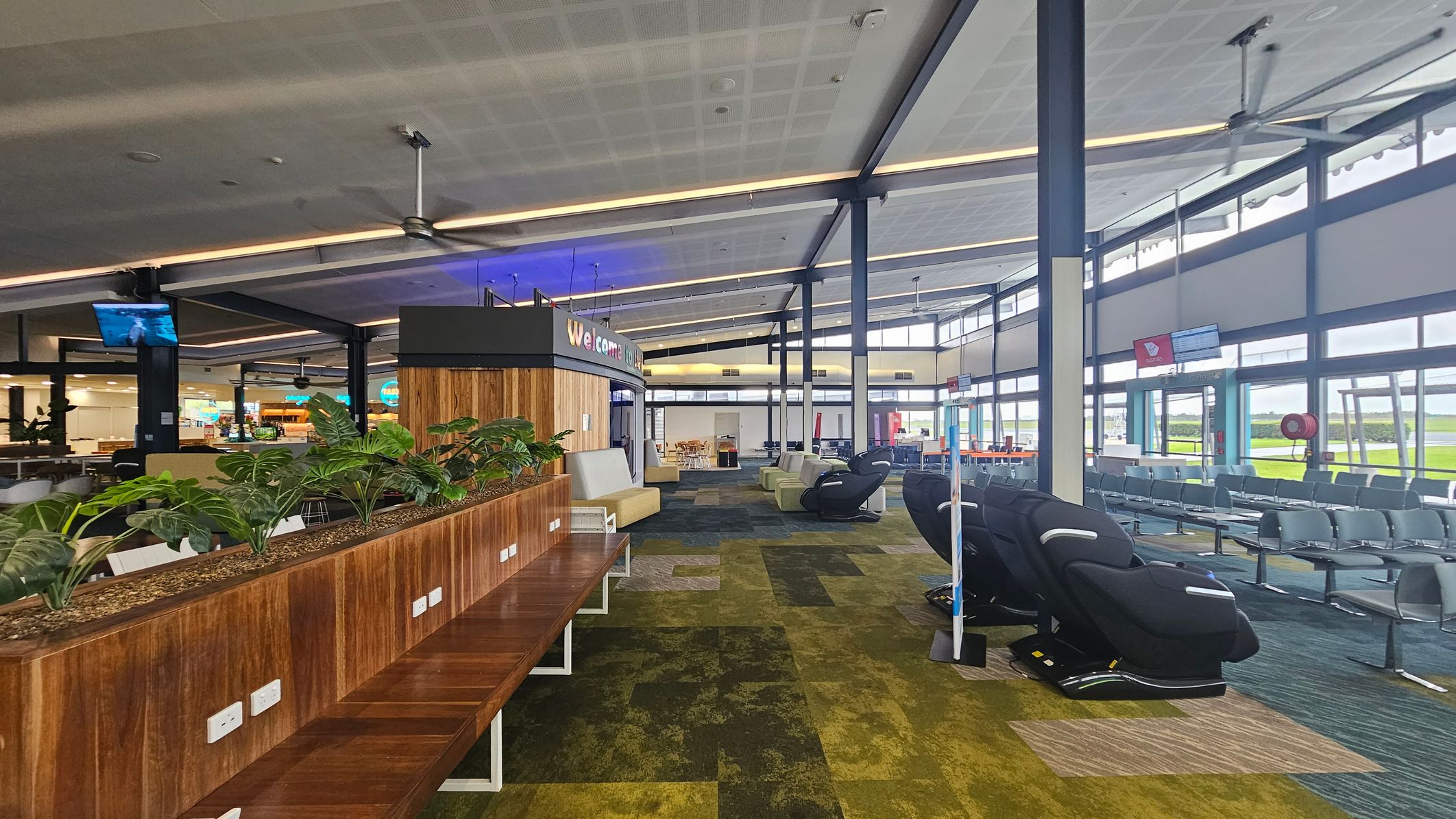 An airport lounge with a modern design. On the left, there's a wooden partition with green plants and power outlets. In the centre, a curved wall with a "Welcome" sign and seating area. On the right, rows of grey chairs and massage chairs. The floor is covered with a patterned carpet in shades of green and brown. The ceiling is high with exposed beams and large fans. Windows on the right let in natural light, offering a view of the tarmac.