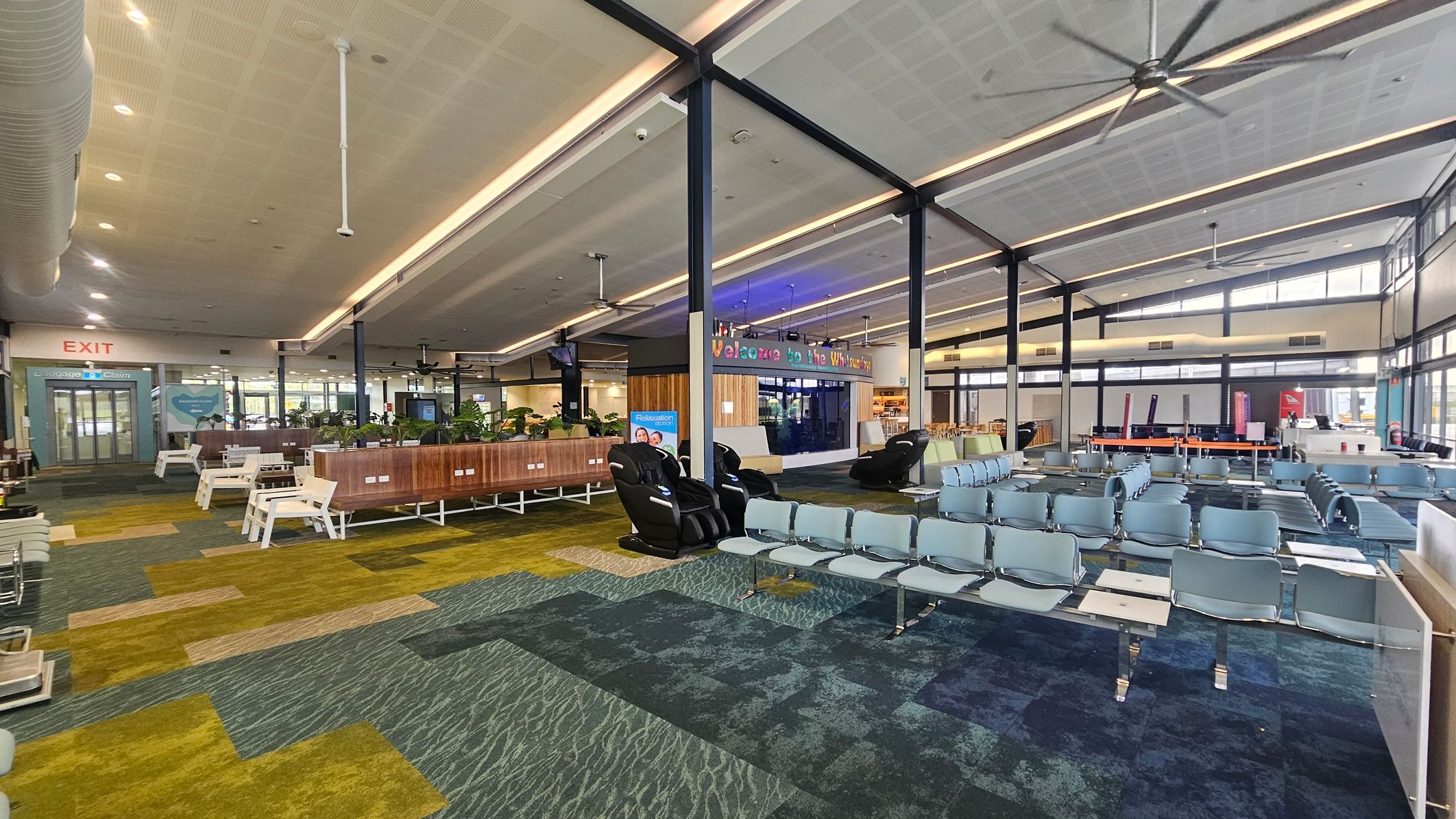 A spacious airport terminal with a patterned carpet in shades of green and blue. On the left, there are white benches and a sign for baggage claim near the exit. In the centre, a wooden partition with plants and massage chairs. On the right, rows of blue seating face a counter area. The ceiling is high with exposed beams and large fans. A colourful sign reads "Welcome to the Whitsundays" above a small shop or café.