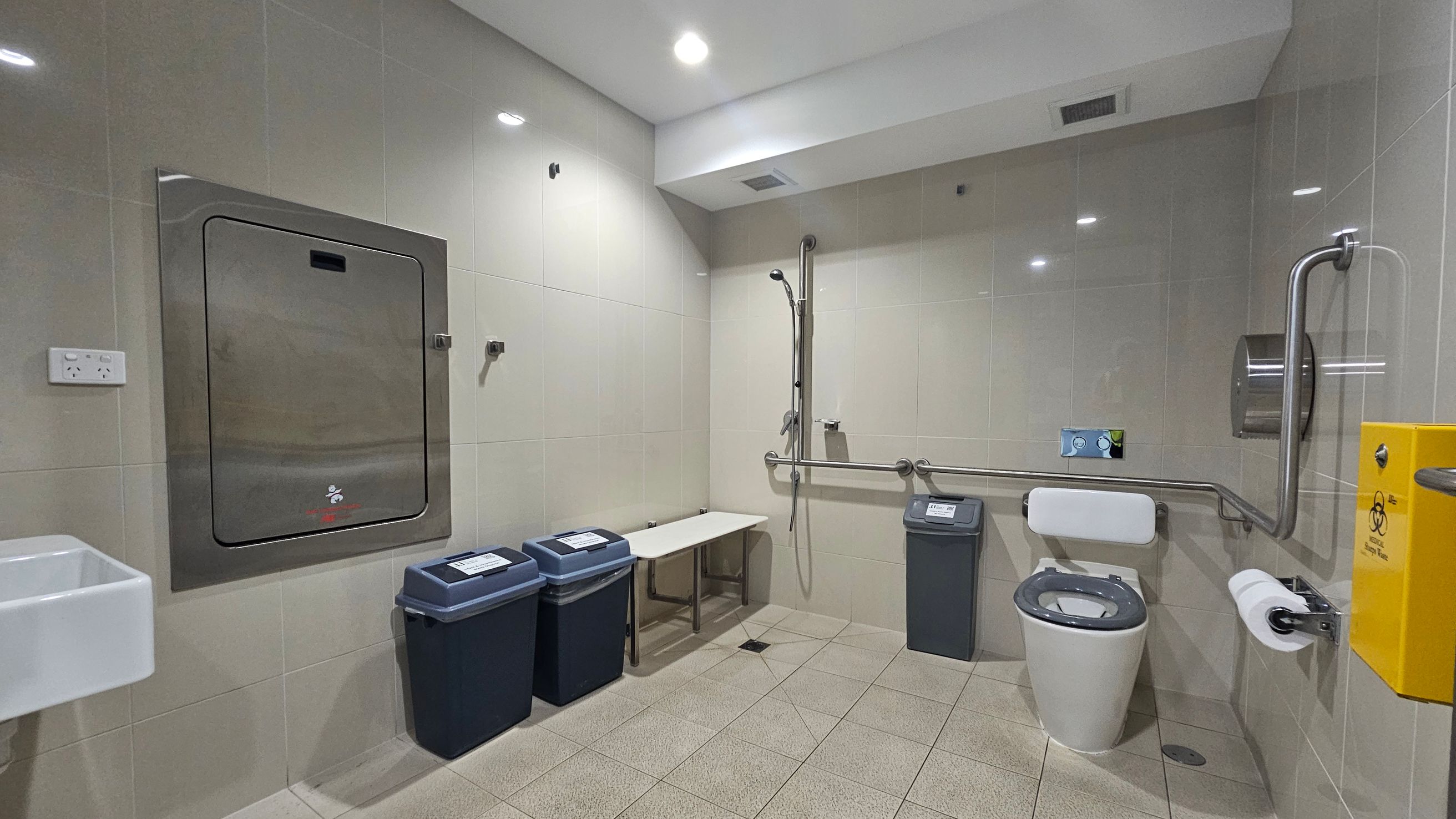 Accessible bathroom with light green tiled walls and floor. On the left, a small white sink and a large metal panel. Below, two bins with blue lids. In the centre, a white bench and a shower with a handheld nozzle and grab bars. On the right, a toilet with support rails, a toilet roll holder, and a yellow biohazard disposal unit. The ceiling is white with recessed lighting.