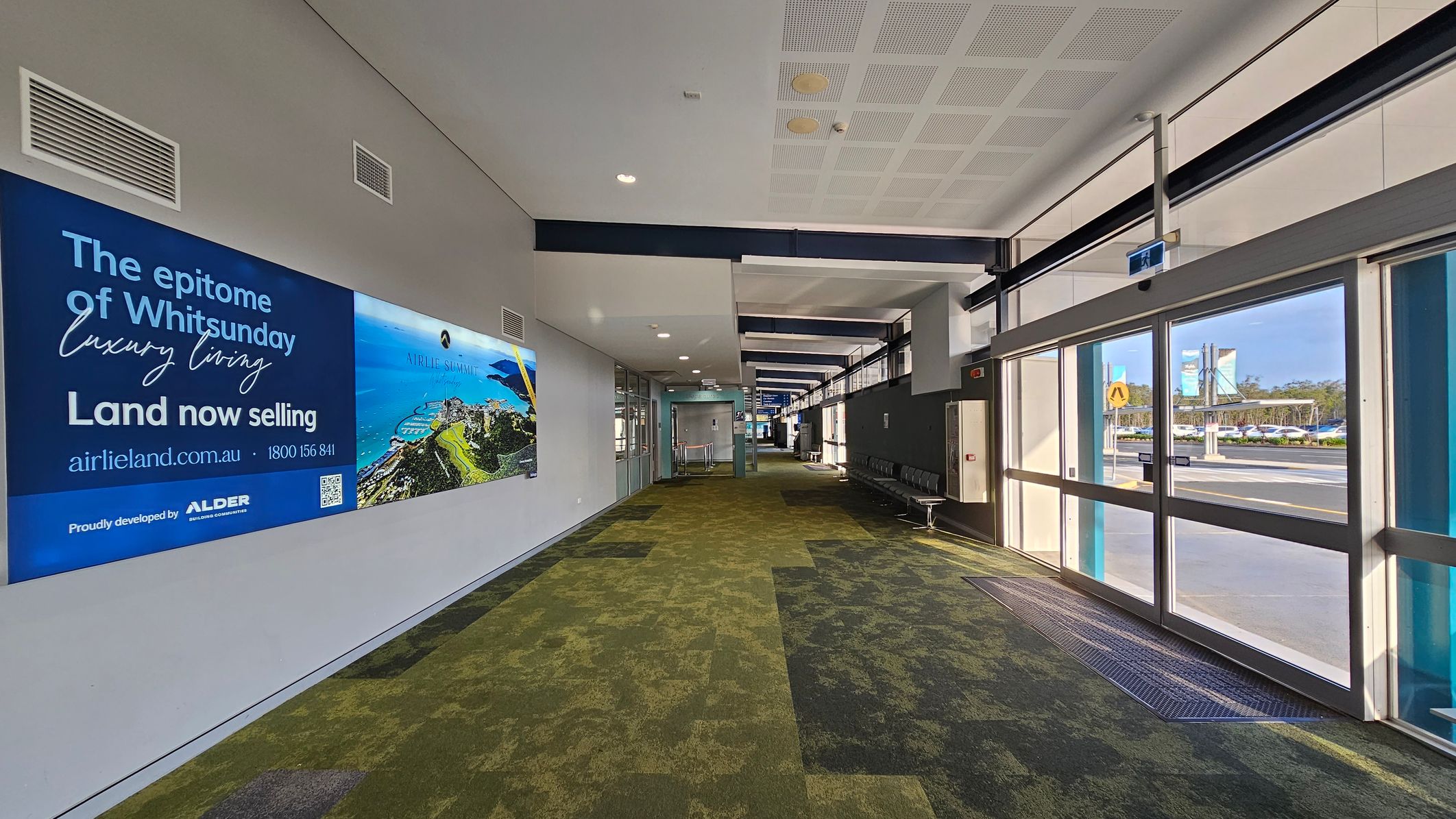 A corridor with a patterned green and brown carpet. On the left, a large advertisement for Whitsunday luxury living is displayed on the wall. The right side features large glass doors leading outside, with a view of a road and parked cars. The ceiling is white with recessed lighting and ventilation panels. Further down the corridor, there are rows of black chairs along the wall.