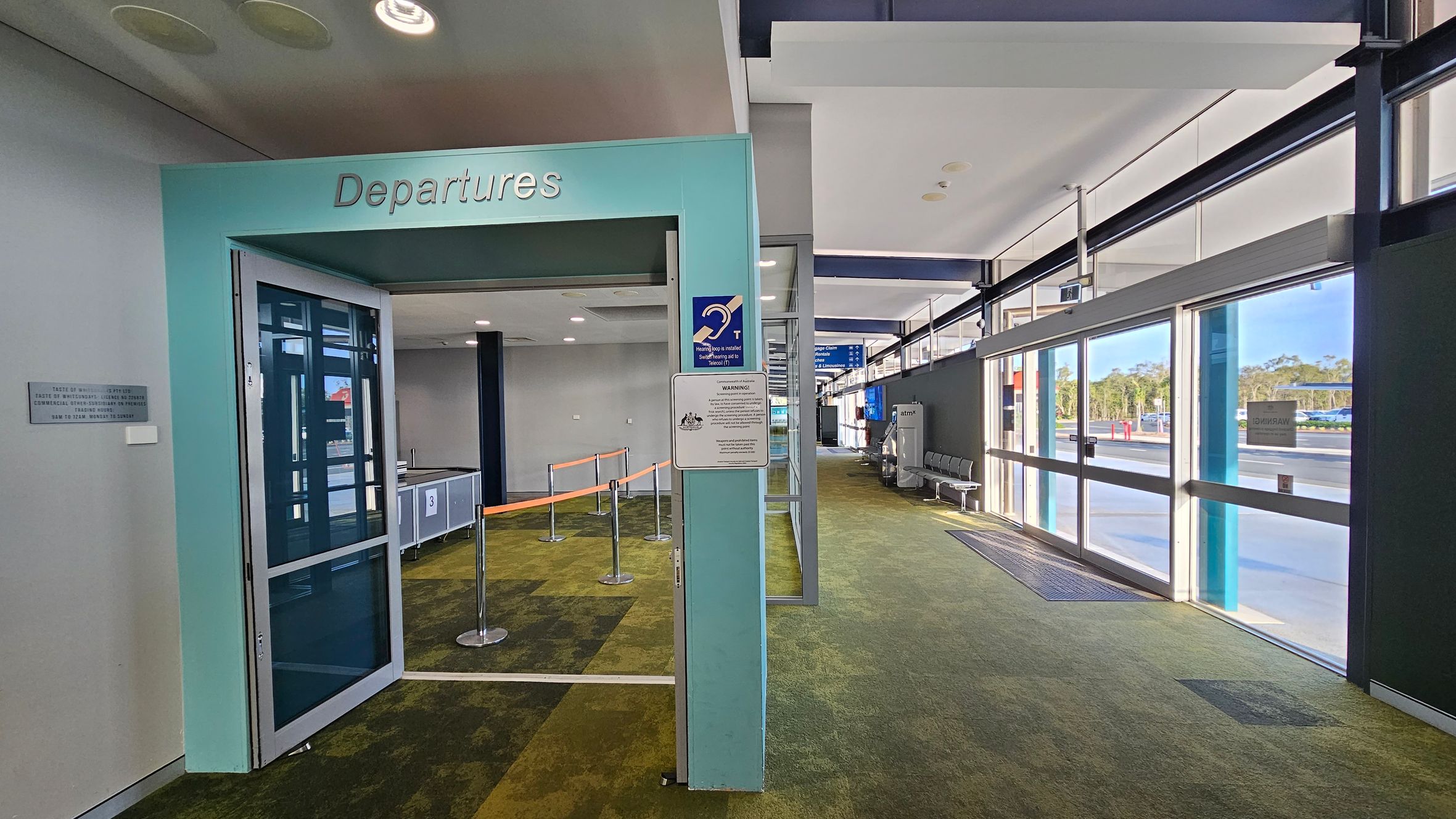 A modern airport interior with a green carpeted floor. On the left, there's a turquoise entrance marked "Departures" with glass doors. Inside, there are orange queue barriers and a security checkpoint. On the right, large glass windows and doors let in natural light, with a view of the outside. Grey seating is visible along the windows. The ceiling is white with recessed lighting.